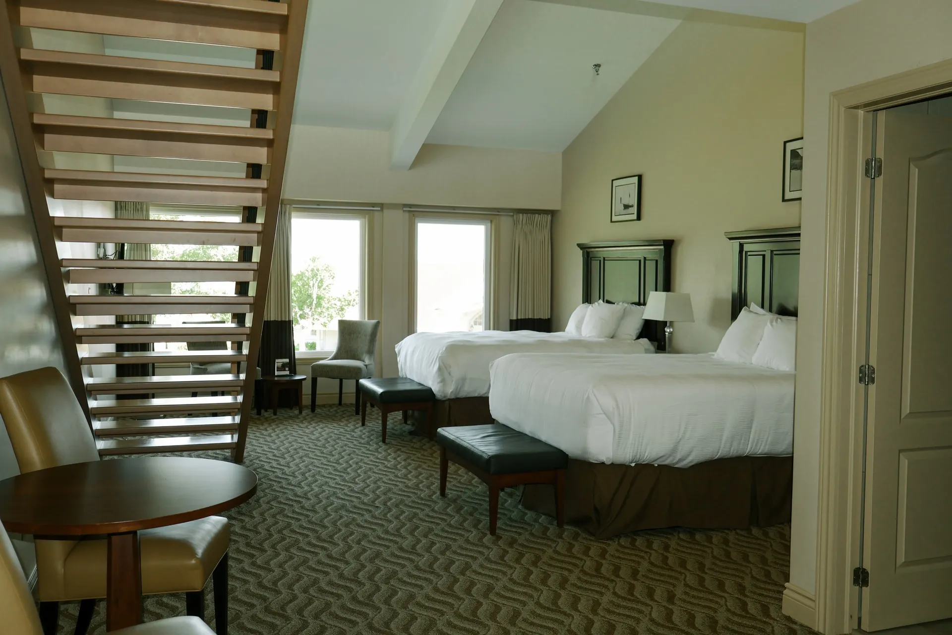A loft suite with two white beds, a wooden staircase, and windows looking onto the grounds.