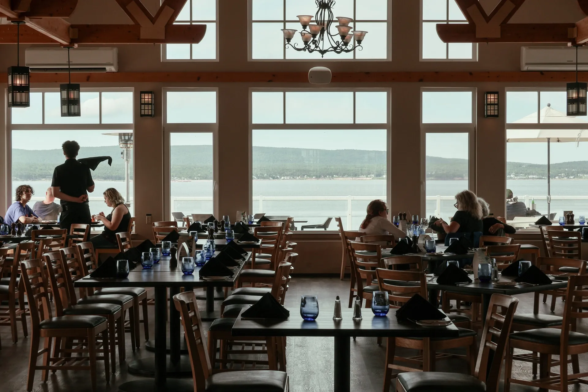 Guests dining beside tall windows at The Arduaine, with the ocean and forested hills beyond.