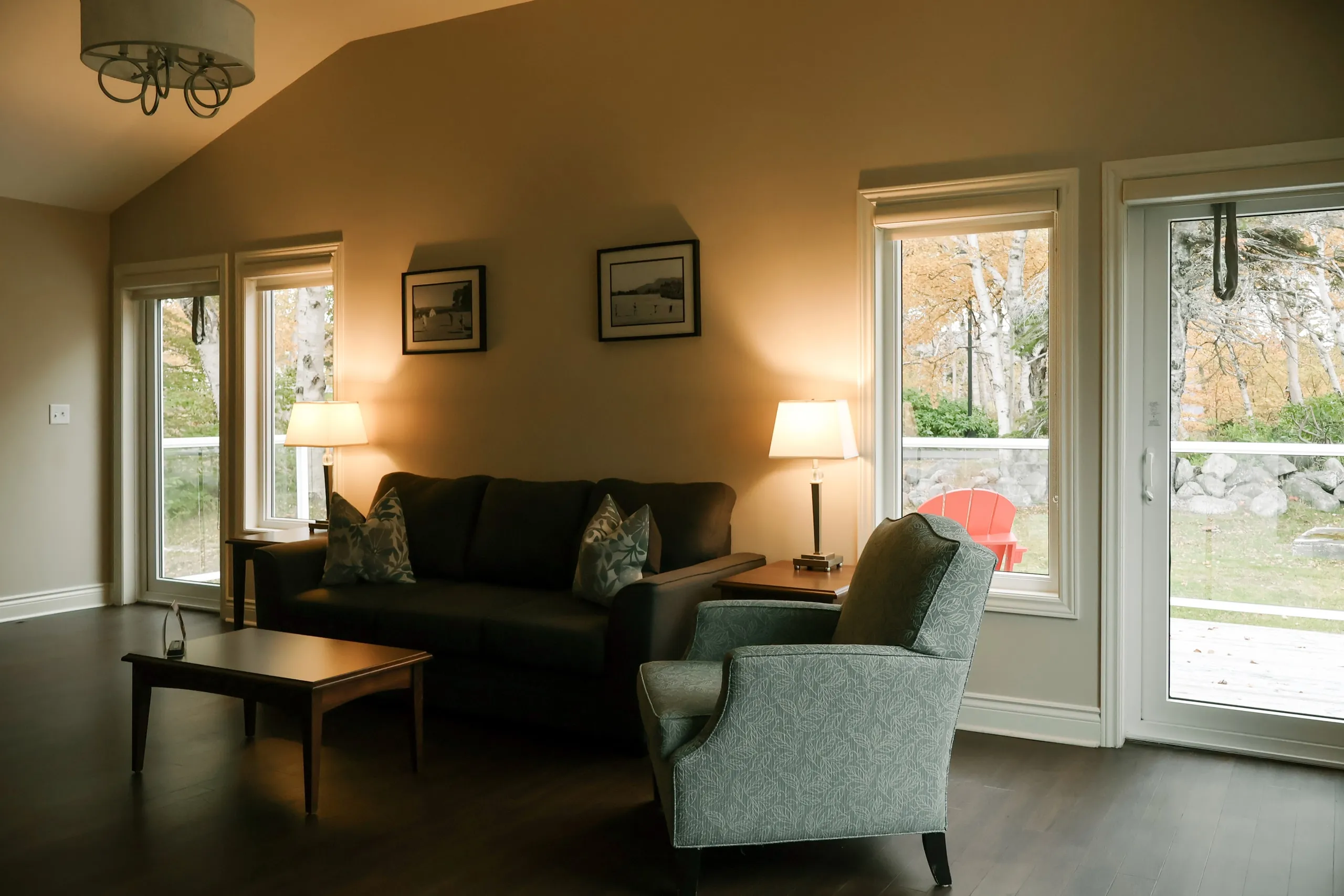 A suite living room with a grey sofa, patterned armchair, wood coffee table, and lamps beside windows to a deck.