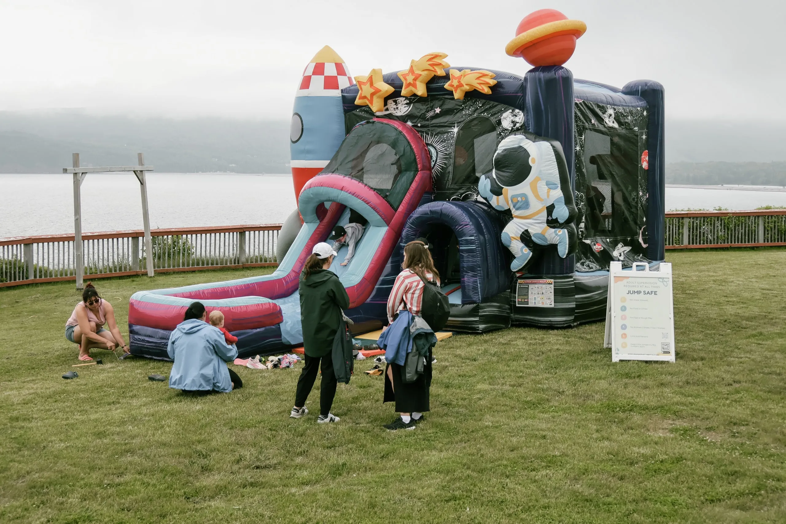 Families gathered on the lawn beside a space-themed bounce house, with the ocean behind a wooden fence.