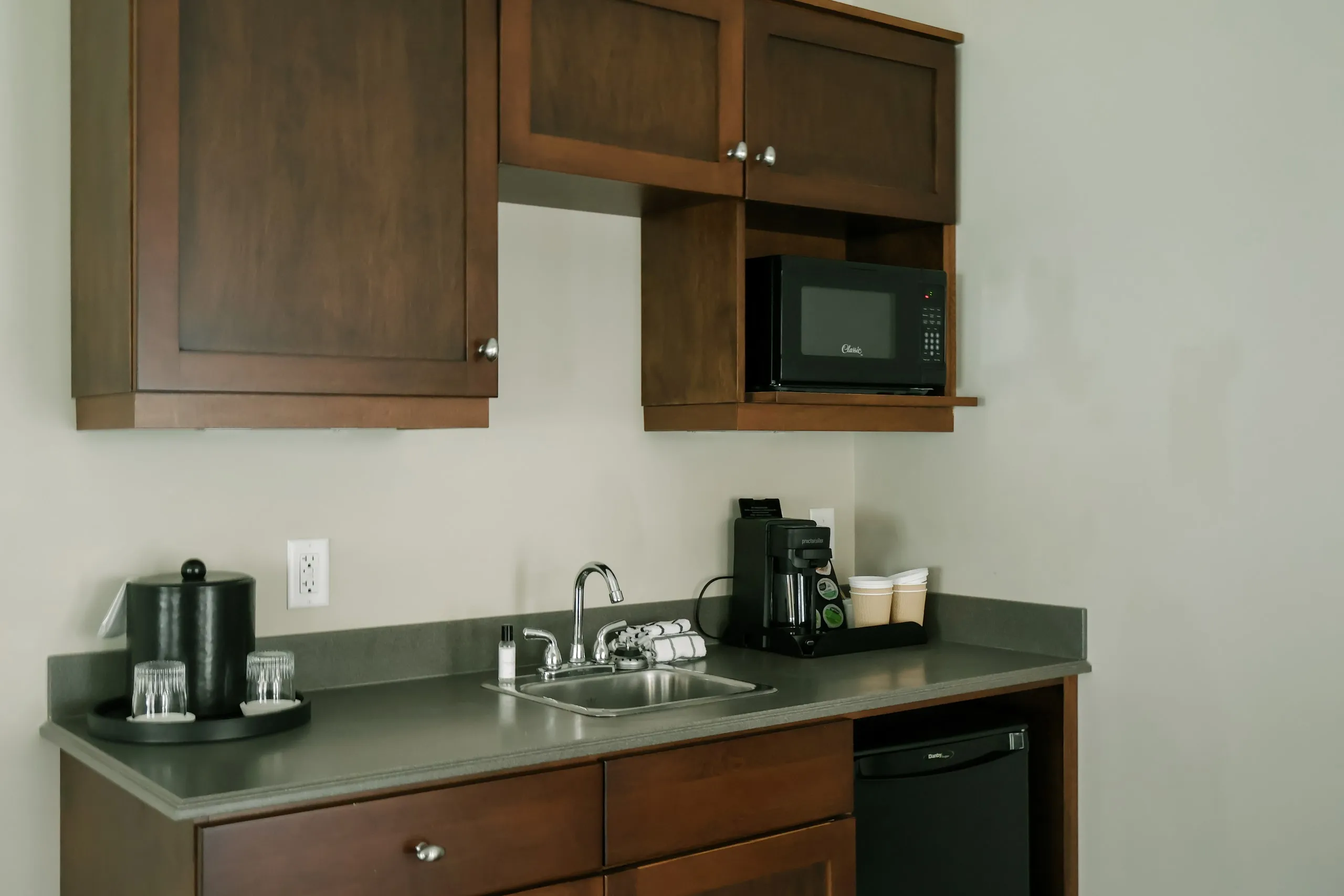 A suite kitchenette with dark wood cabinets, a grey countertop, microwave, mini fridge, and a coffee station.