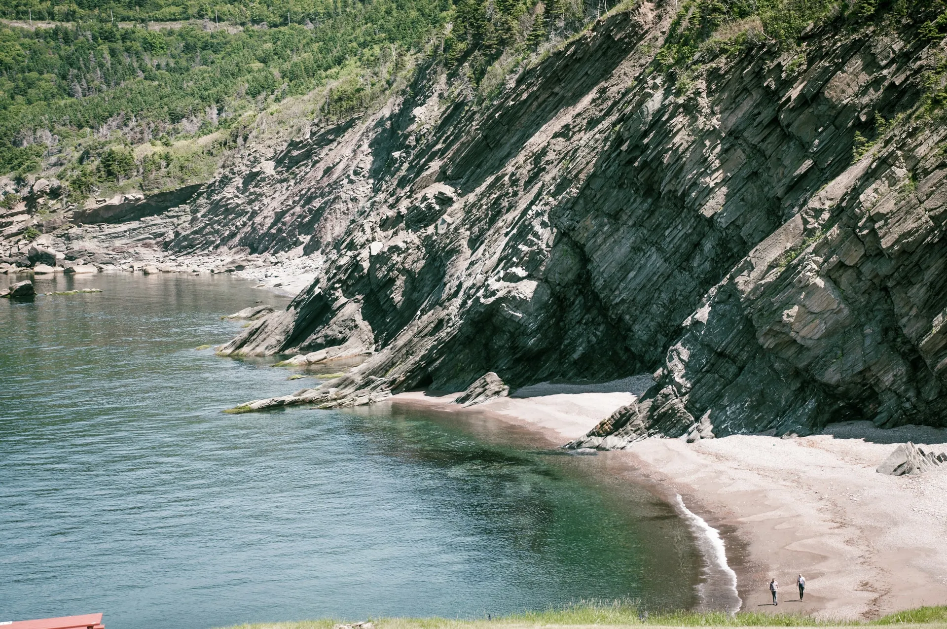 Two people on a sandy cove beneath steep layered cliffs along the Atlantic.