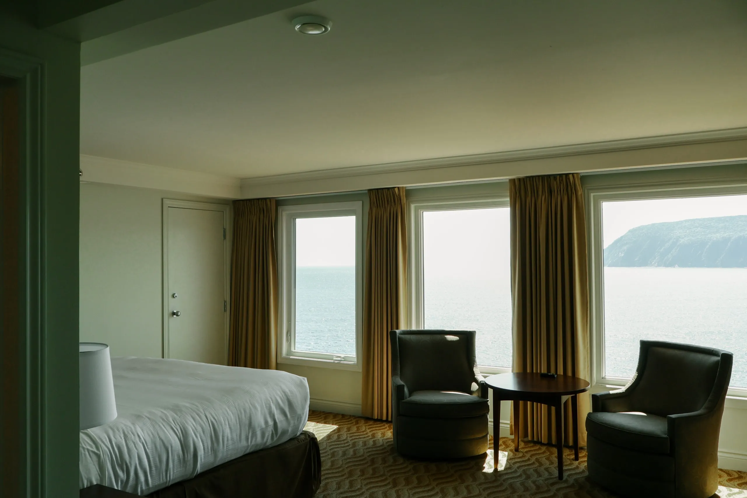 A guest room with two beds, gold curtains, and armchairs facing windows over the water.