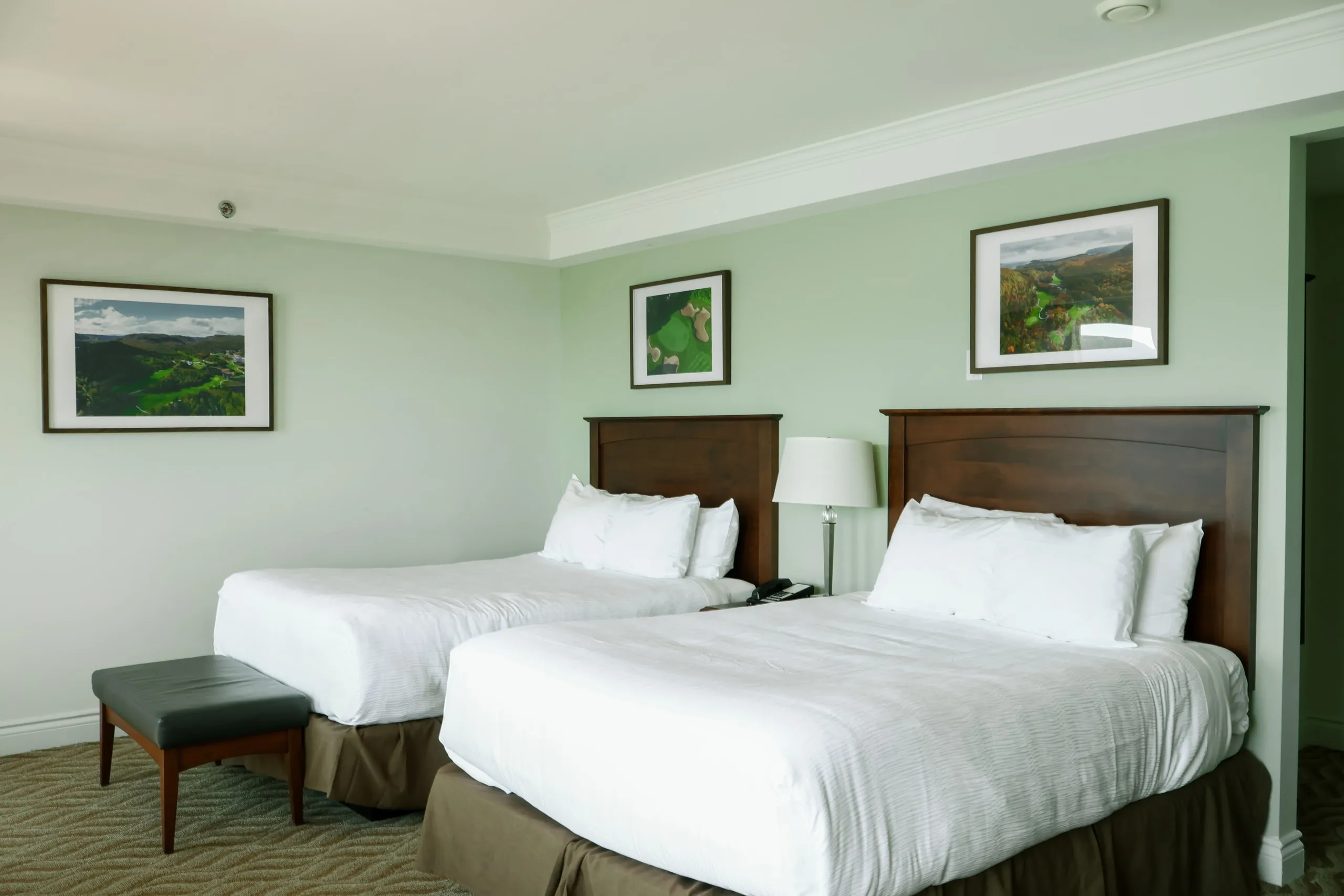 A guest room with two white beds, dark wood headboards, and framed highland landscapes on pale green walls.