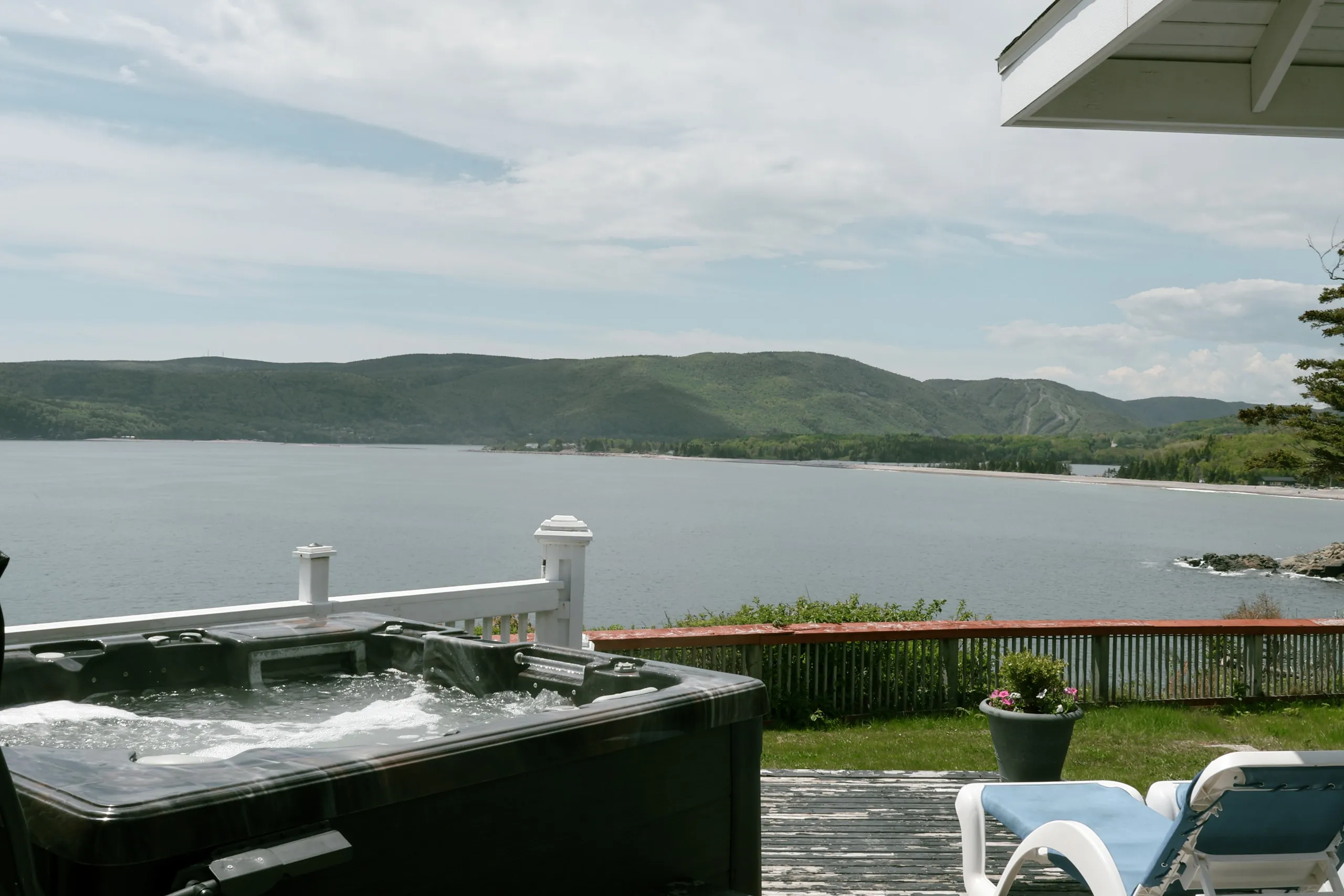 A hot tub on a deck beside lounge chairs, looking across the water to forested hills.