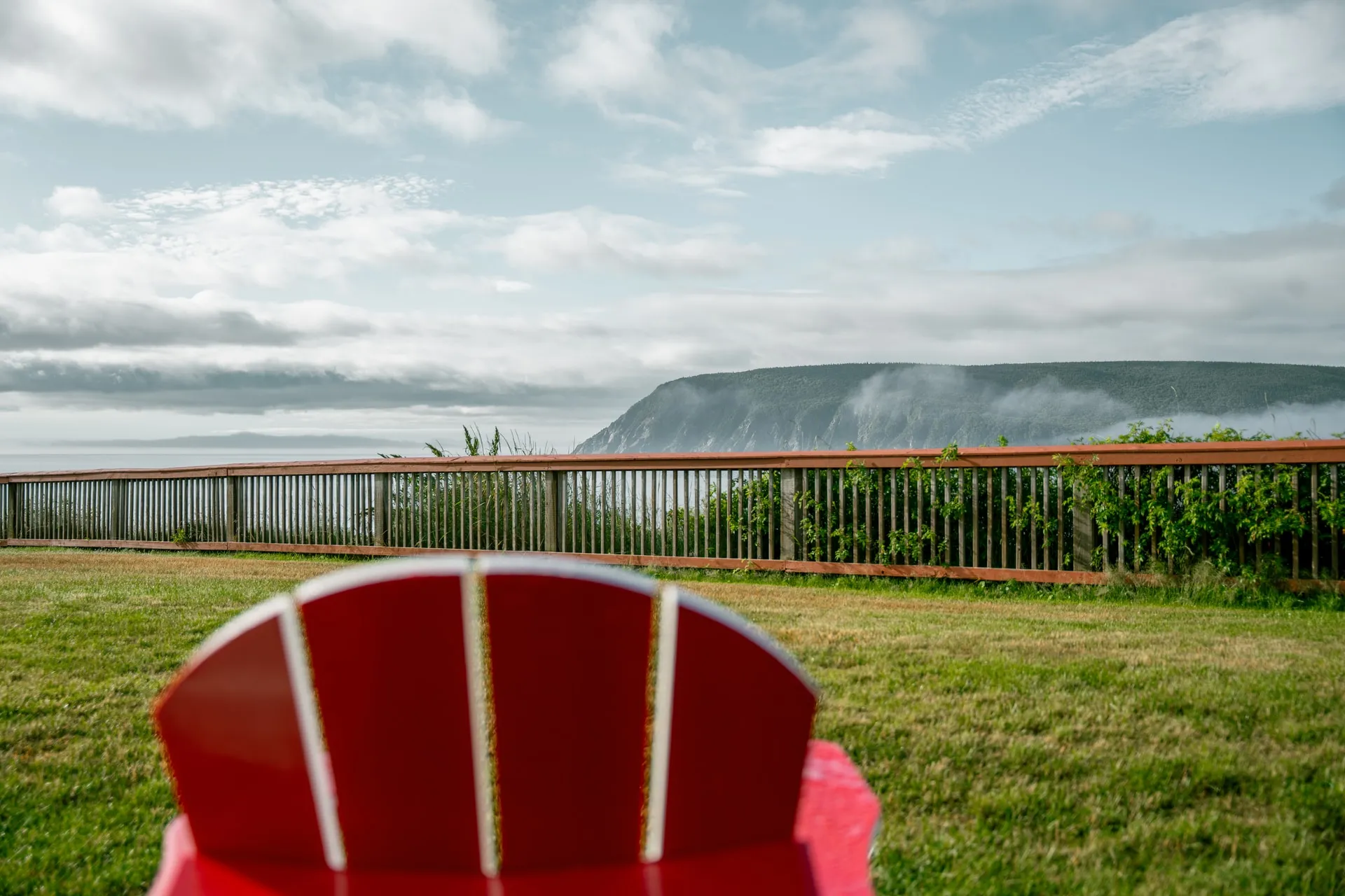 A red Adirondack chair on the lawn facing a wooden fence, the ocean, and a misty cliff.