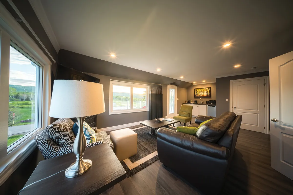A living area with a dark leather sofa, patterned armchair, cream ottoman, and a lamp on a desk beside the windows.
