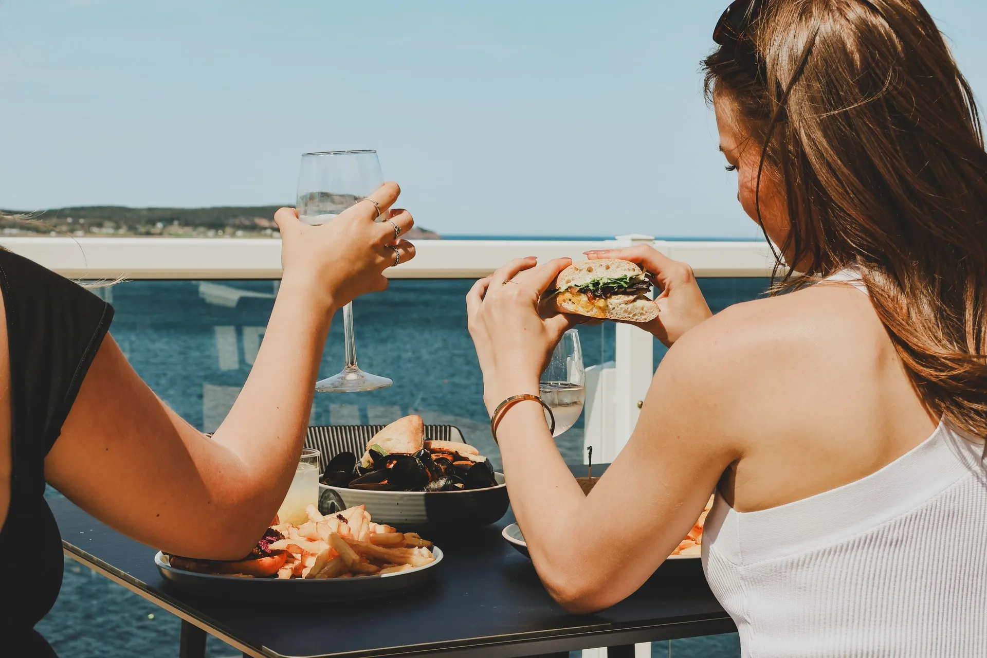 Two guests sharing wine and a meal on an oceanfront patio, with the water beyond the railing.
