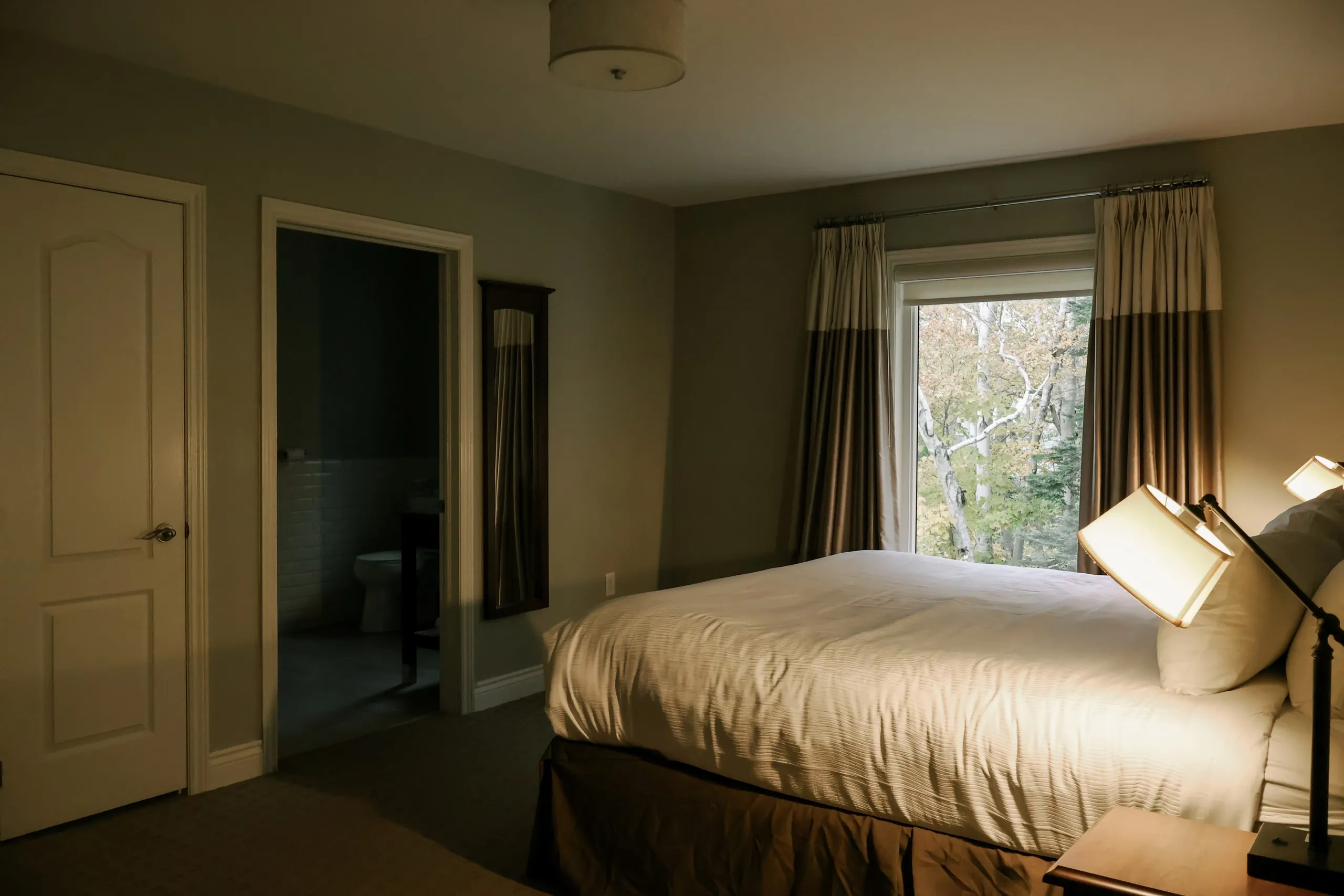 A king bedroom with white linens, an open doorway to the ensuite bathroom, and autumn trees outside the window.