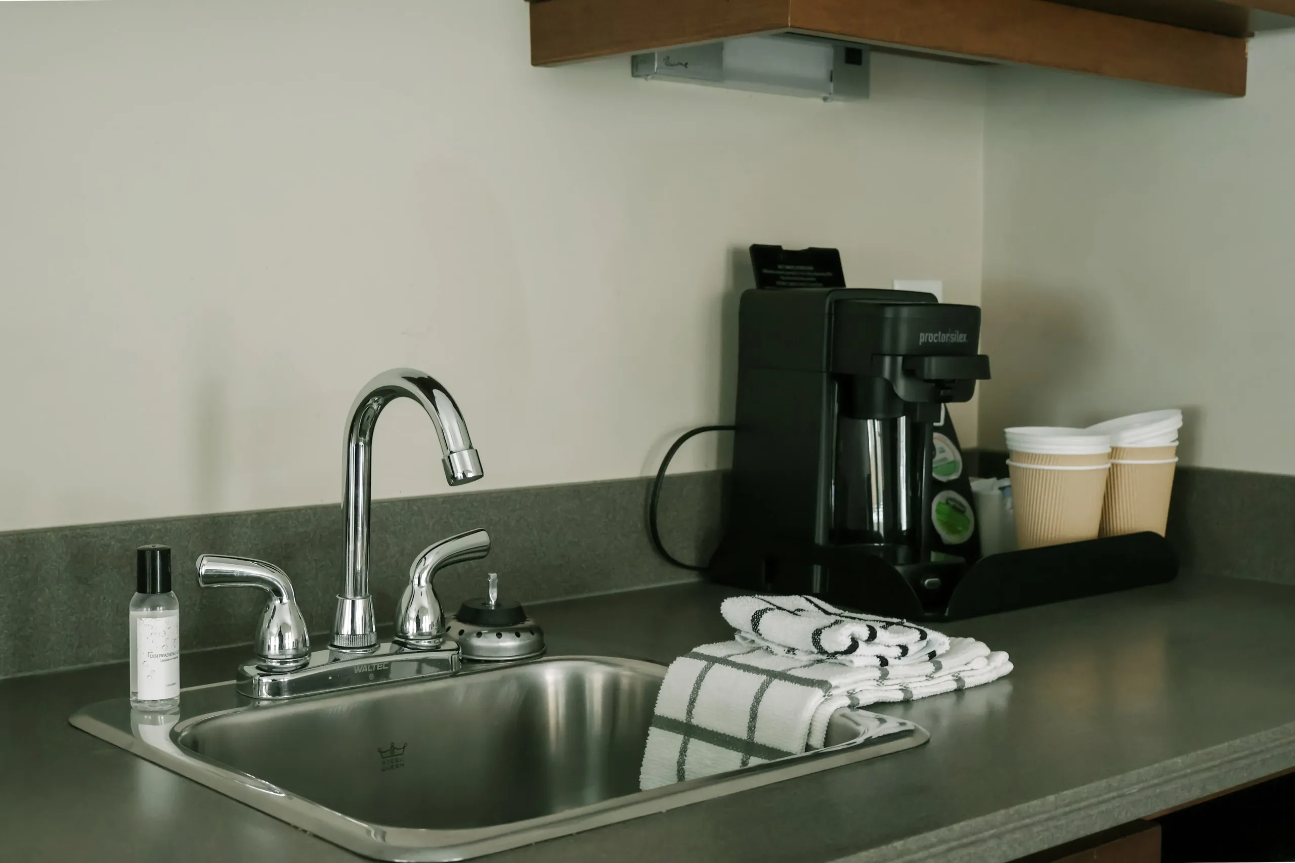 A kitchenette countertop with a stainless sink, chrome faucet, coffee maker, and stacked paper cups.