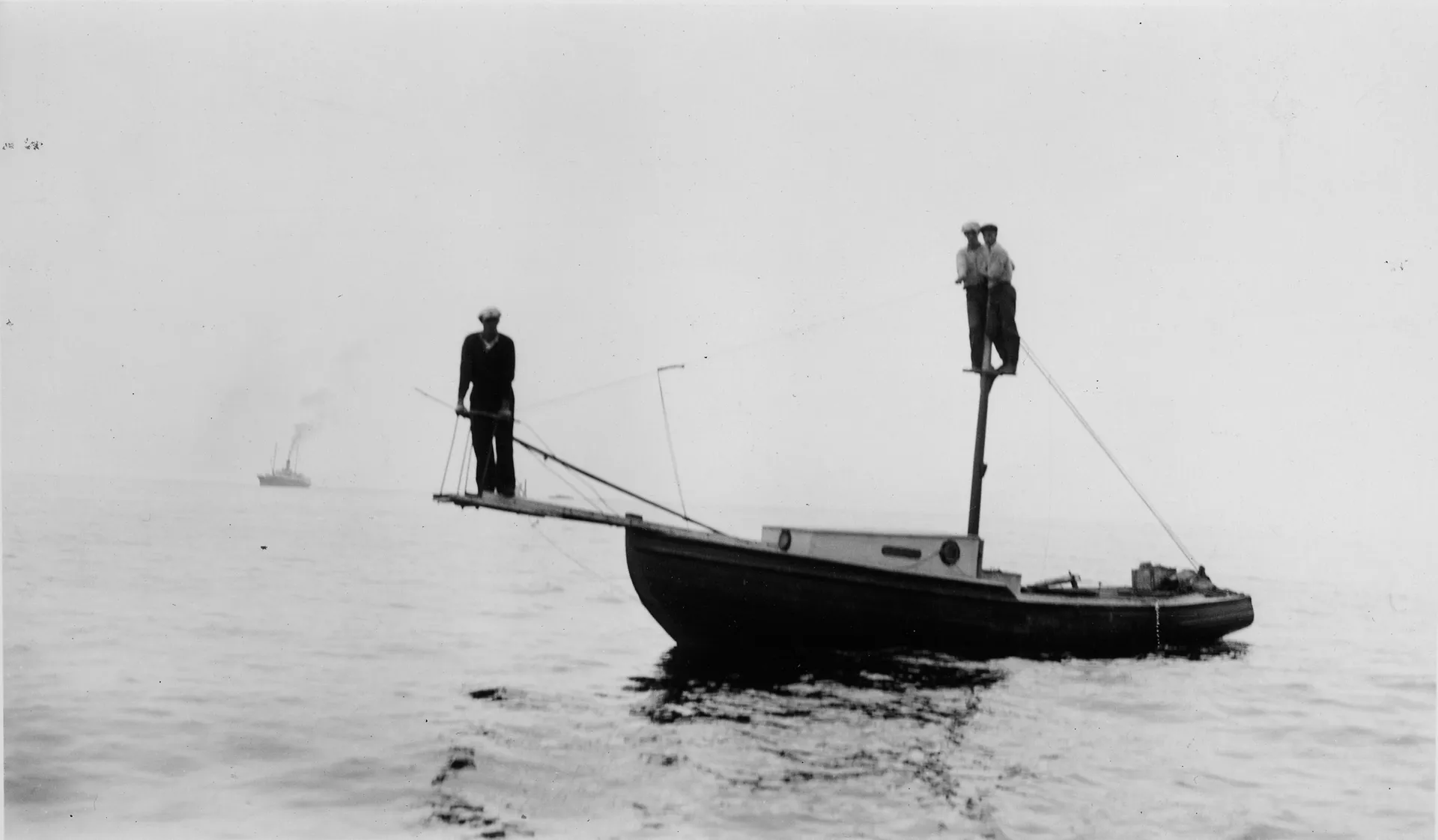 A man stands on a long wooden plank extending from the bow of a boat in a black-and-white archival photograph.