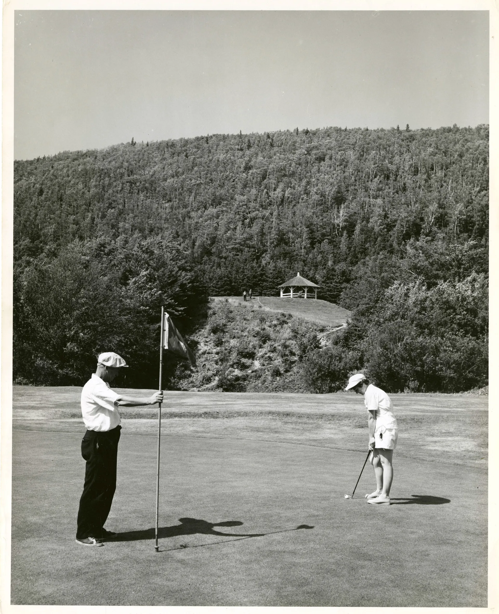 Two men in vintage golf attire on a green, one putting, in a black-and-white archival photograph.