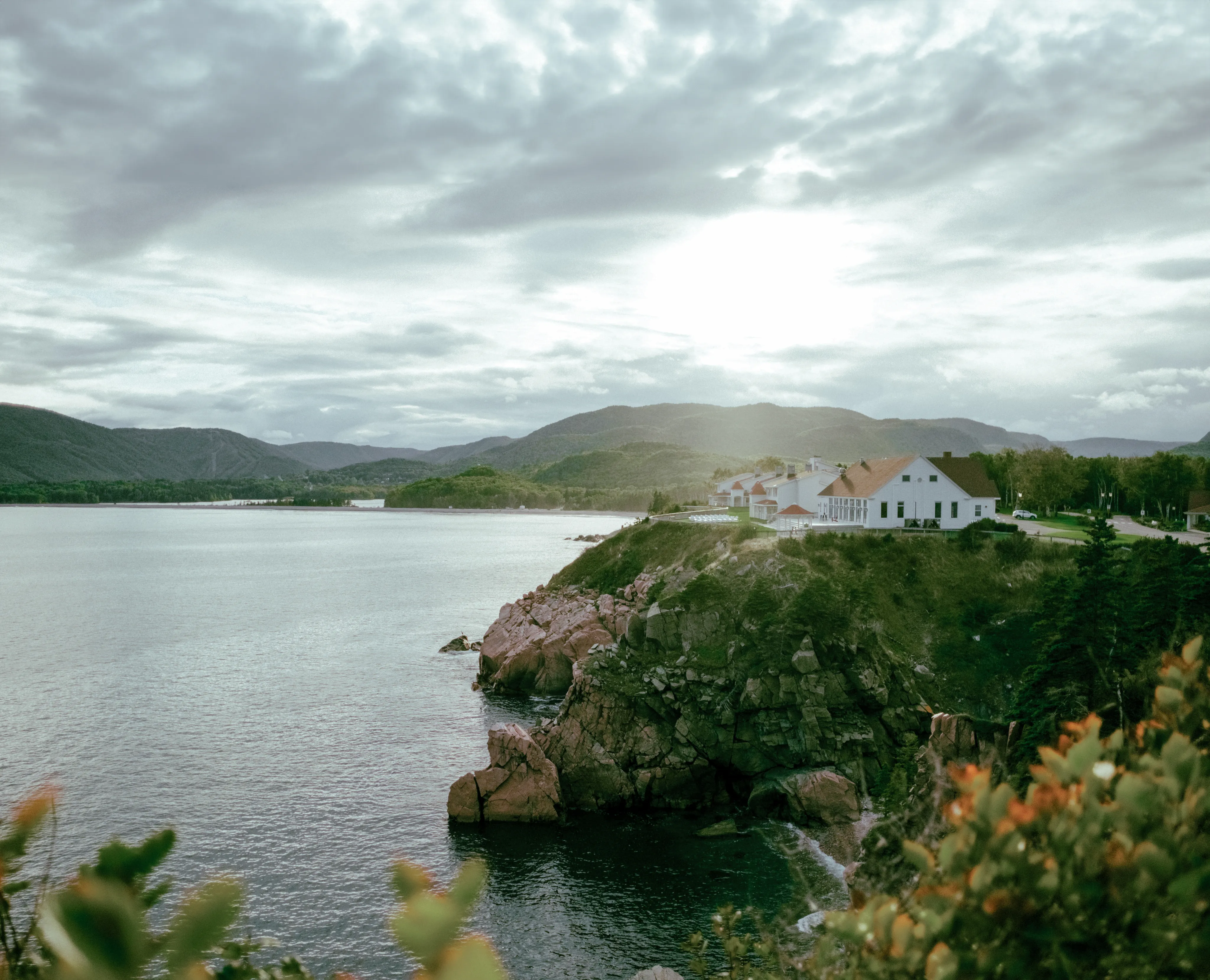 The white Keltic Lodge on a rocky headland above the ocean, with forested mountains under a cloudy sky.