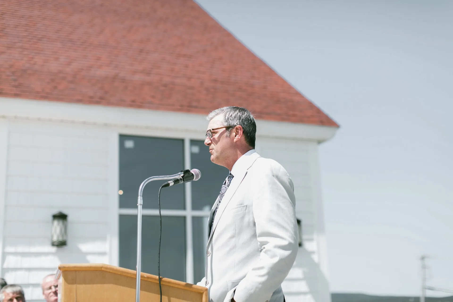 A speaker at a podium in front of a white building with a red roof.