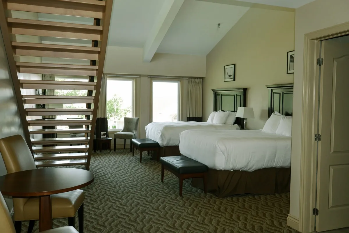 A loft guest room with an open wooden staircase, two beds, and windows along the far wall.