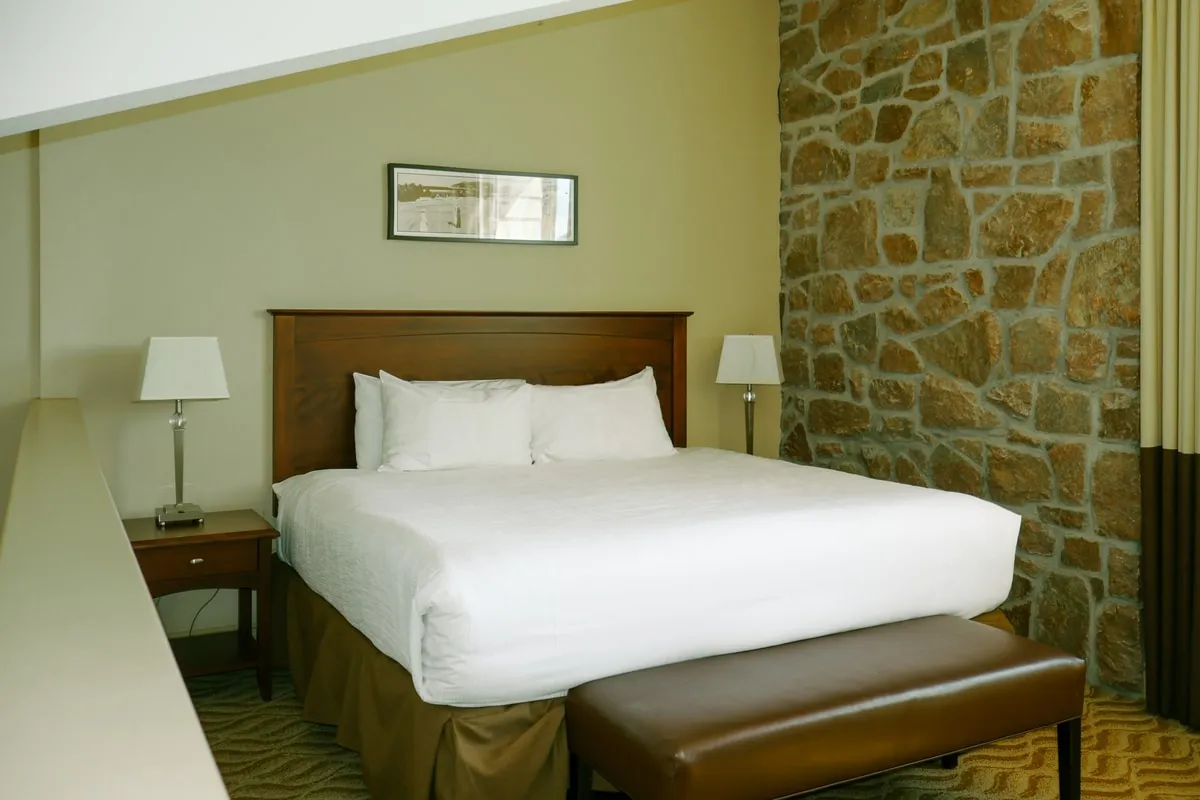A loft bedroom with a white bed beside a stone wall and a bench at the foot of the bed.