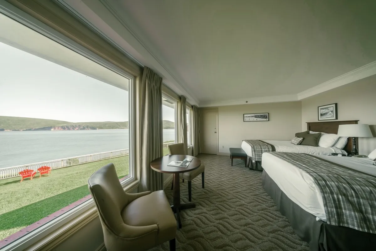 A guest room with two beds and a table for two beside windows looking onto the lawn and the water.