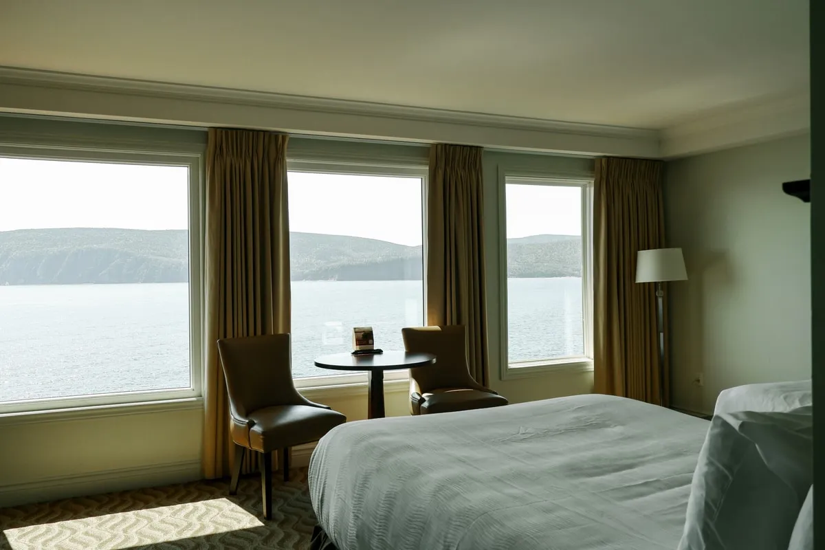 A guest room with a bed, a round table, and two chairs beside windows overlooking the water and the hills.