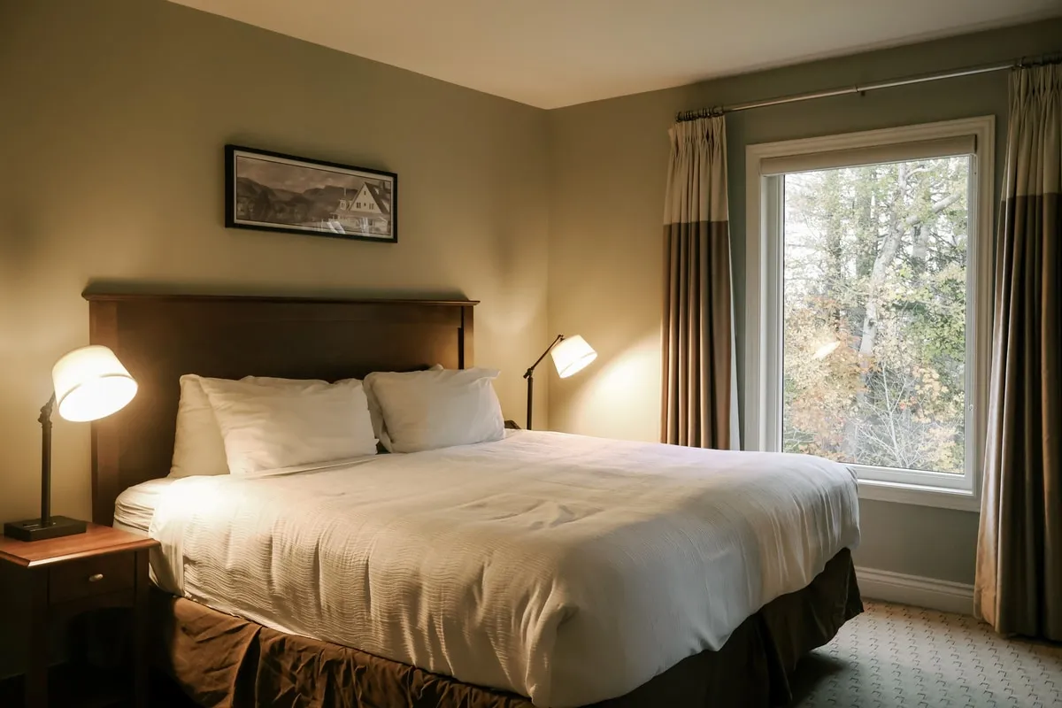 A king bedroom with a dark wood headboard, a framed landscape above the bed, and a window looking onto the trees.