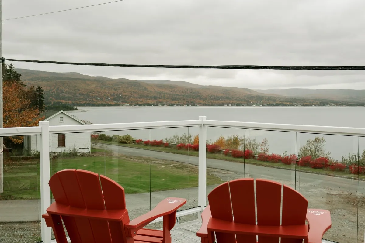 Two red Adirondack chairs on a wooden deck with a glass railing, looking across the water toward autumn hills.