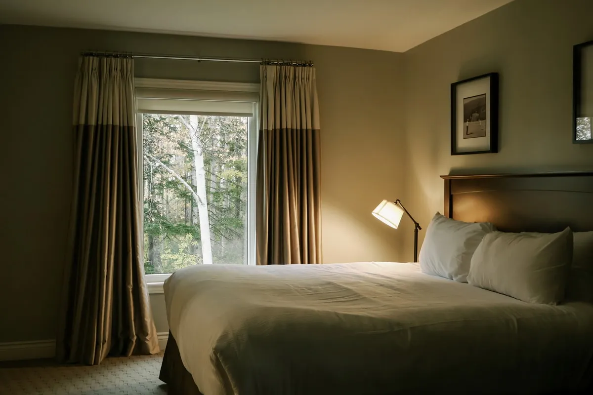 A king bedroom with a wood headboard, white linens, and two-tone curtains framing a window onto the trees.