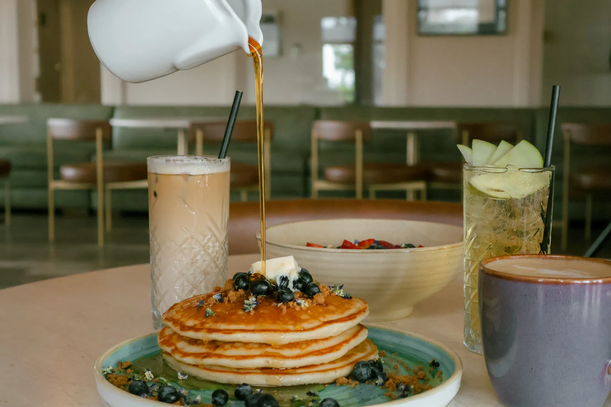 Syrup poured over a stack of pancakes with berries and butter, beside an iced latte, a fruit drink, and a coffee.