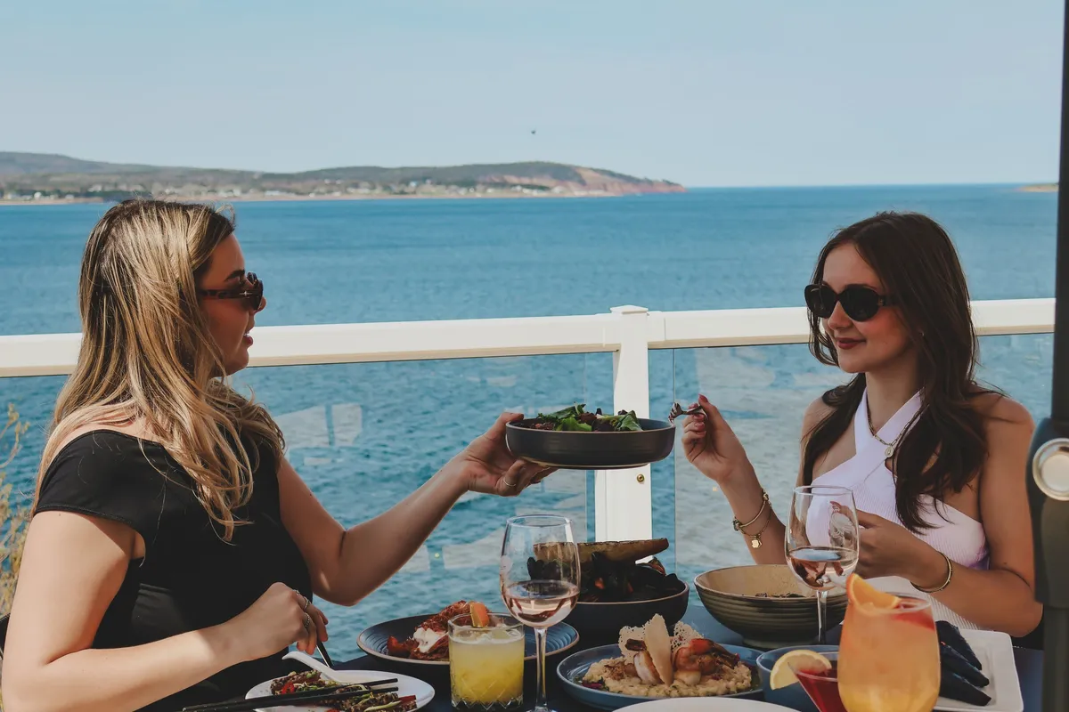 Two guests sharing a salad at an oceanfront table with the Atlantic and coastline behind them.