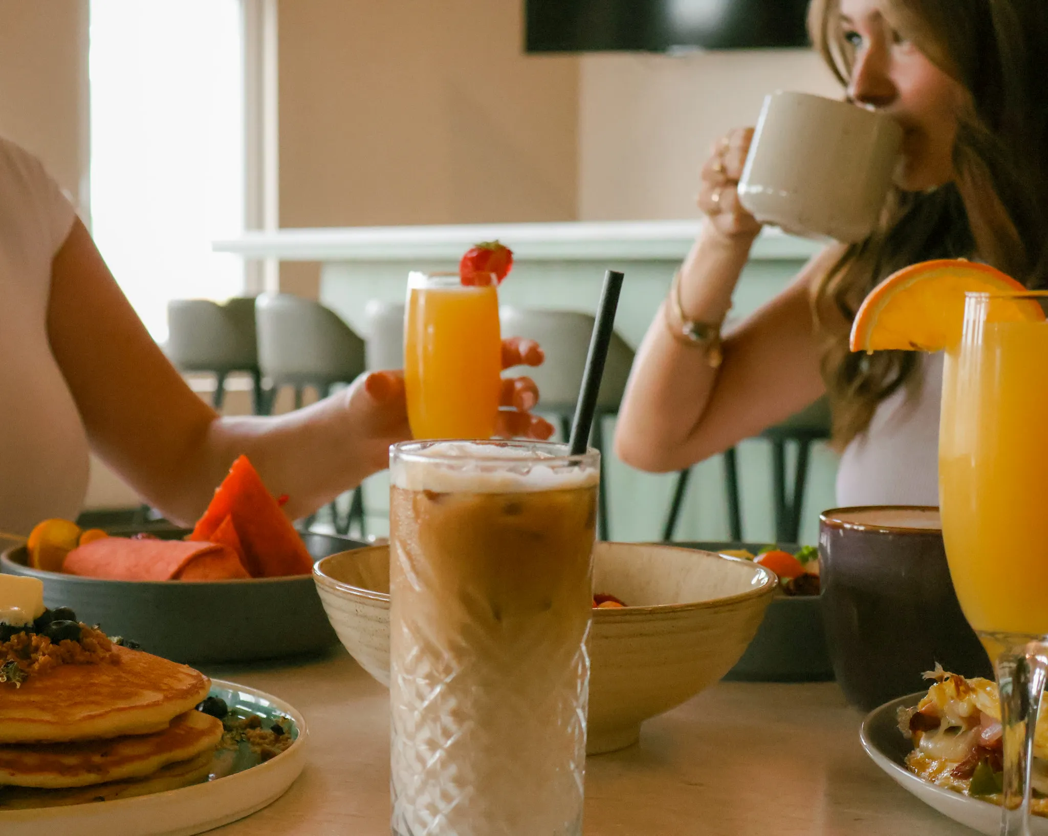 Two people at a breakfast table, one sipping coffee beside glasses of orange juice, an iced latte, and pancakes.
