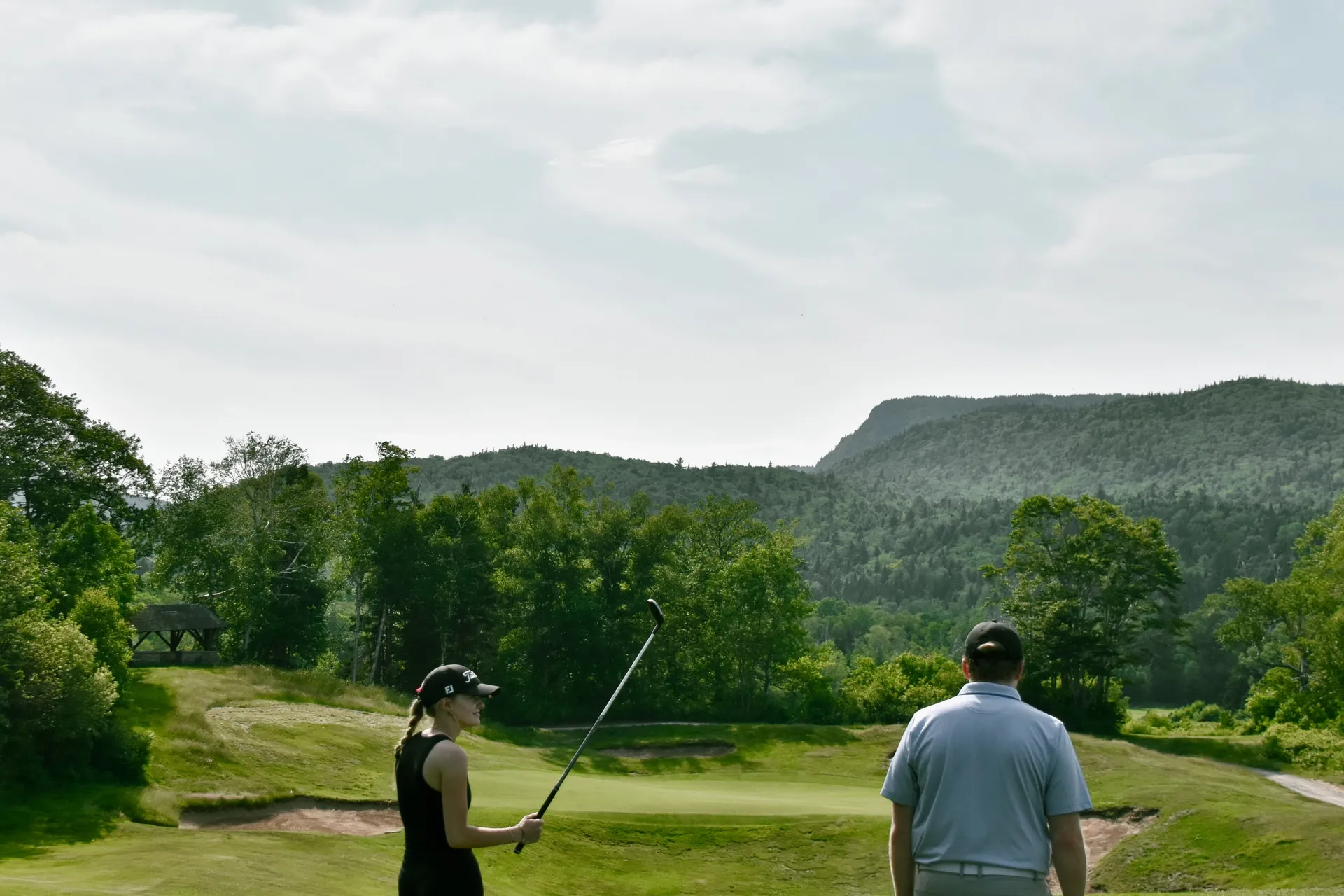 A golfer at the top of her backswing beside a bunker, with a partner watching and a forested mountain beyond.