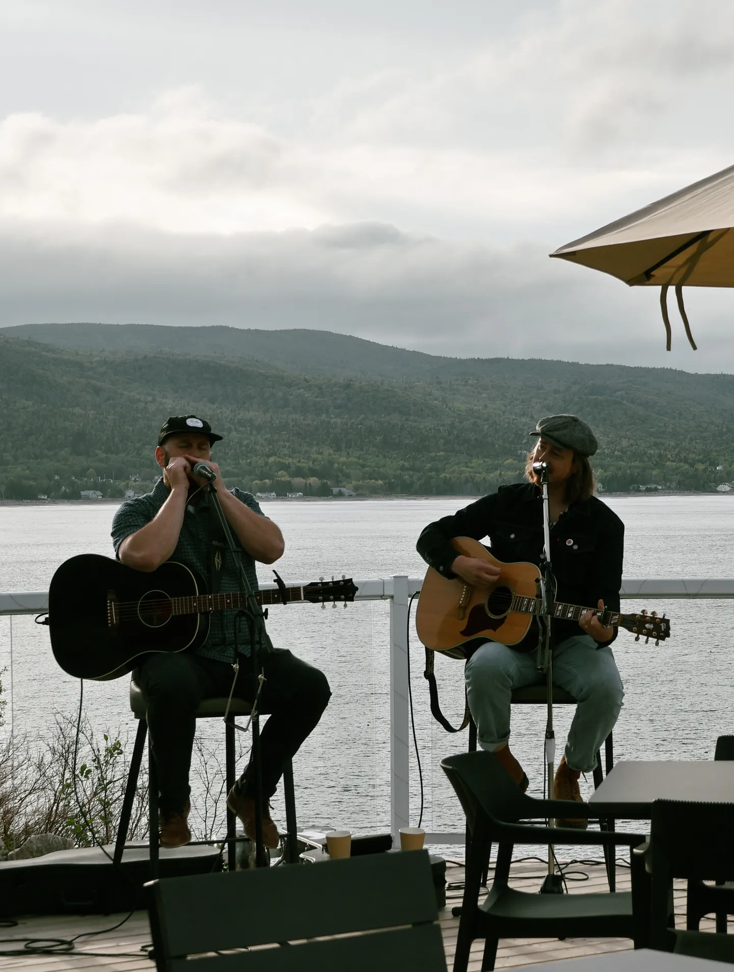 Two musicians performing with guitars on an oceanfront patio, forested hills across the water.
