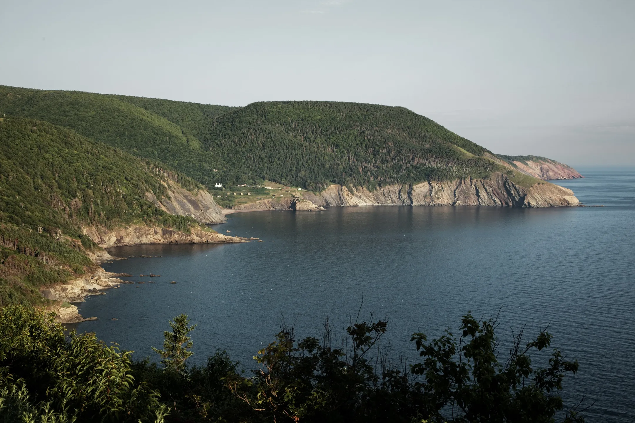 Forested hills and rocky coastline along a blue bay in the Cape Breton Highlands.