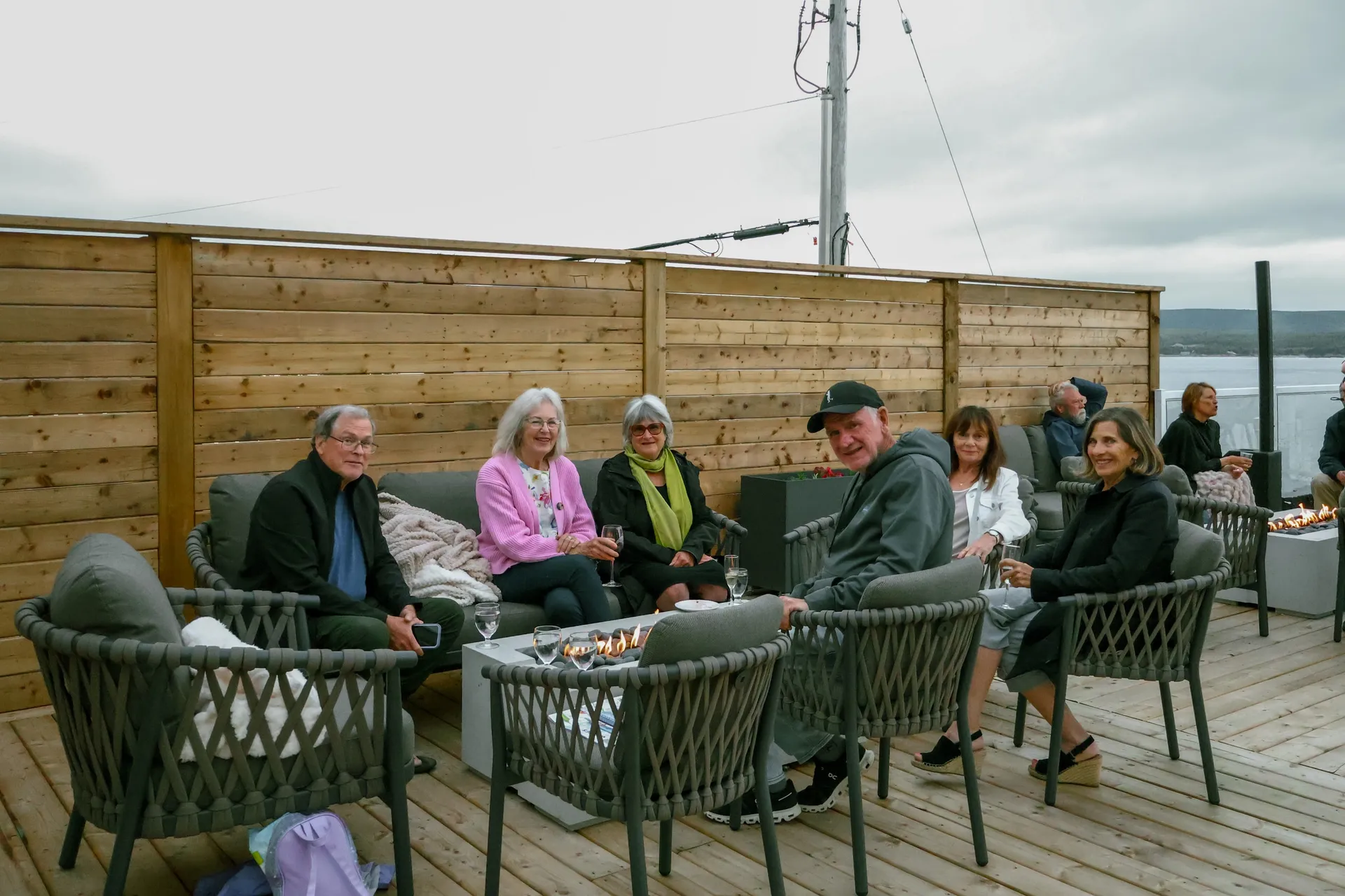 Guests gathered around a fire pit on a wooden deck overlooking the water.