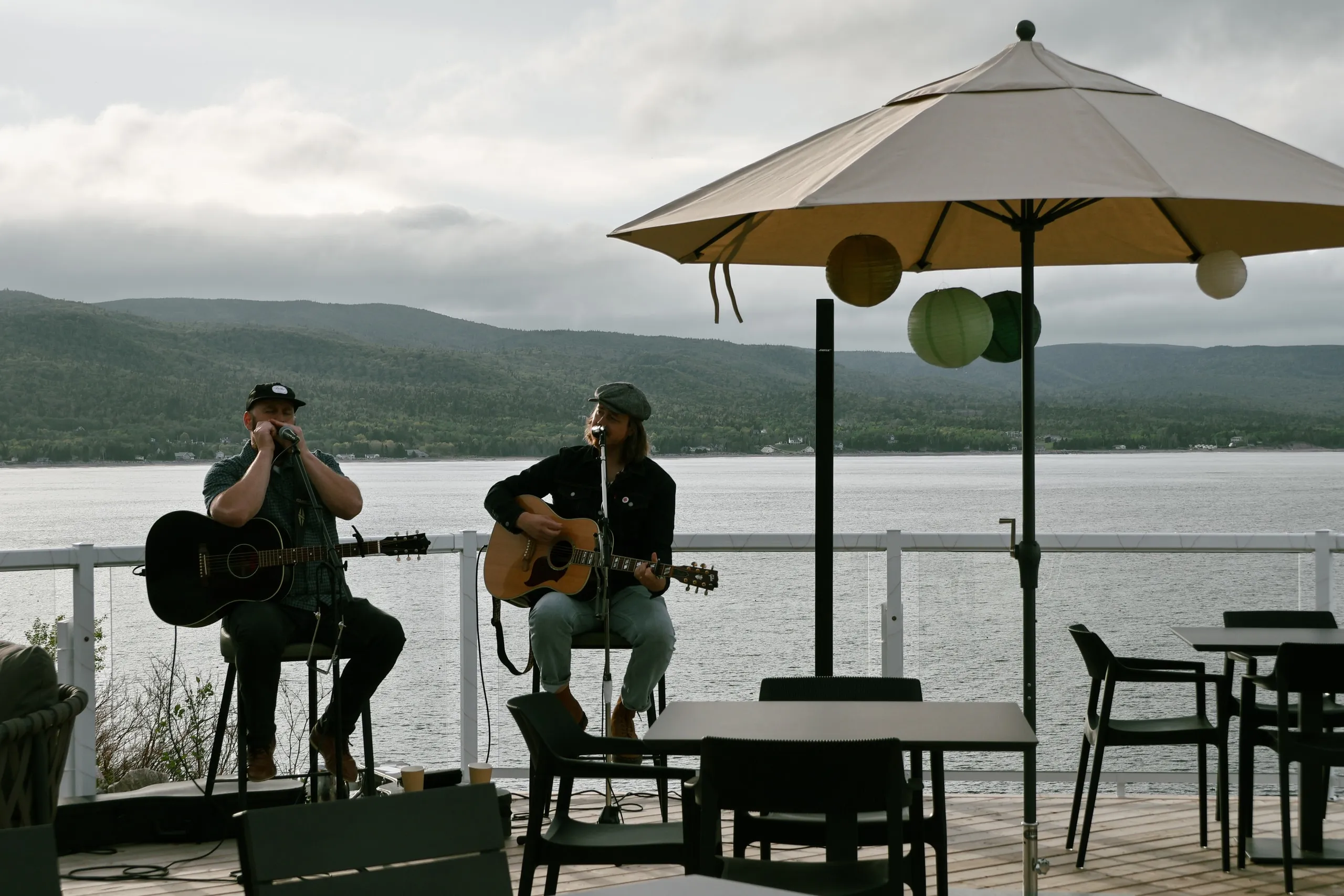 Musicians playing guitars under a white umbrella on the oceanfront patio, with the Atlantic and hills behind them.