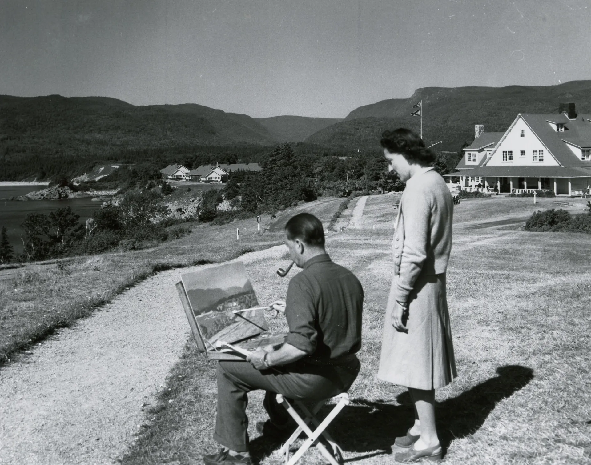 A historical photograph of a man painting at an easel on the lawn while a woman watches, with the lodge and the Highlands behind them.