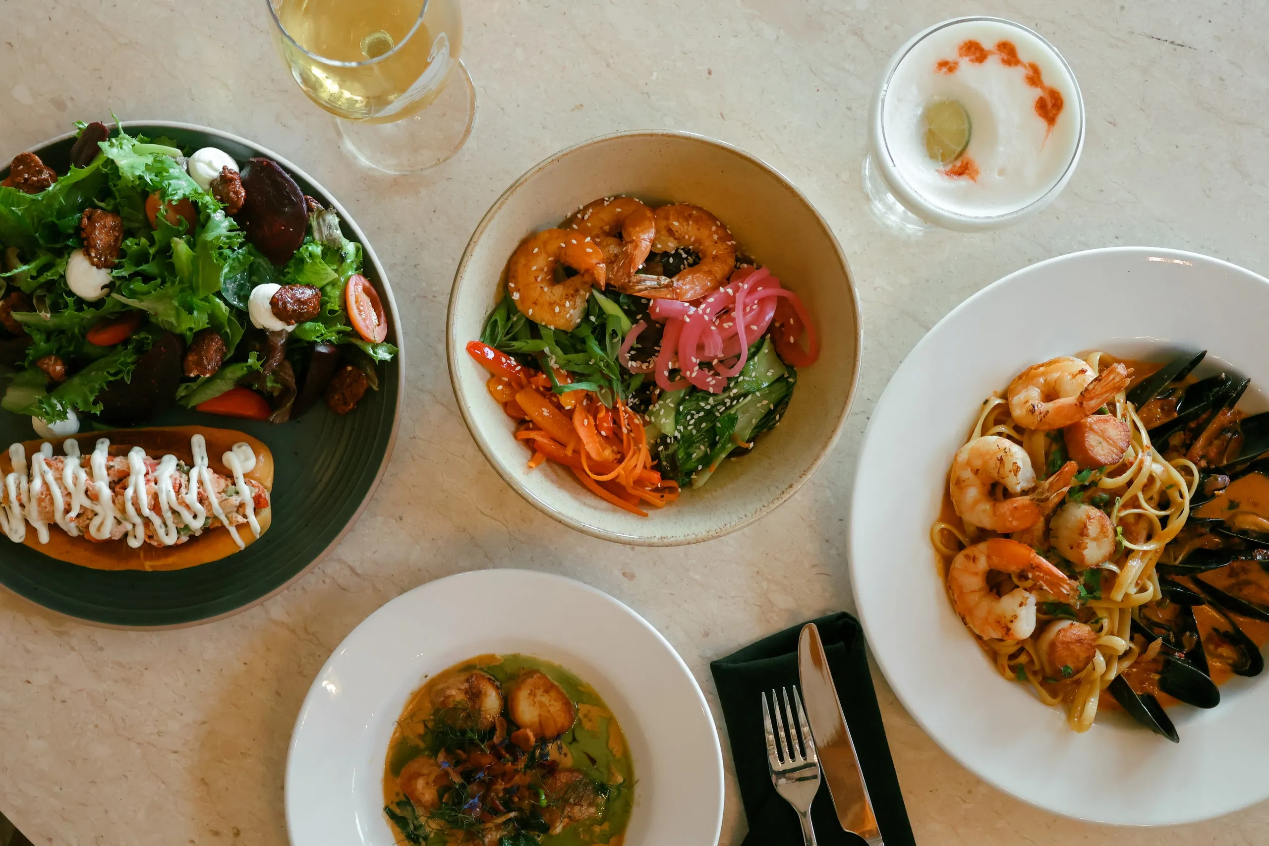An overhead spread of lobster roll, shrimp bowl, scallops, and seafood pasta.