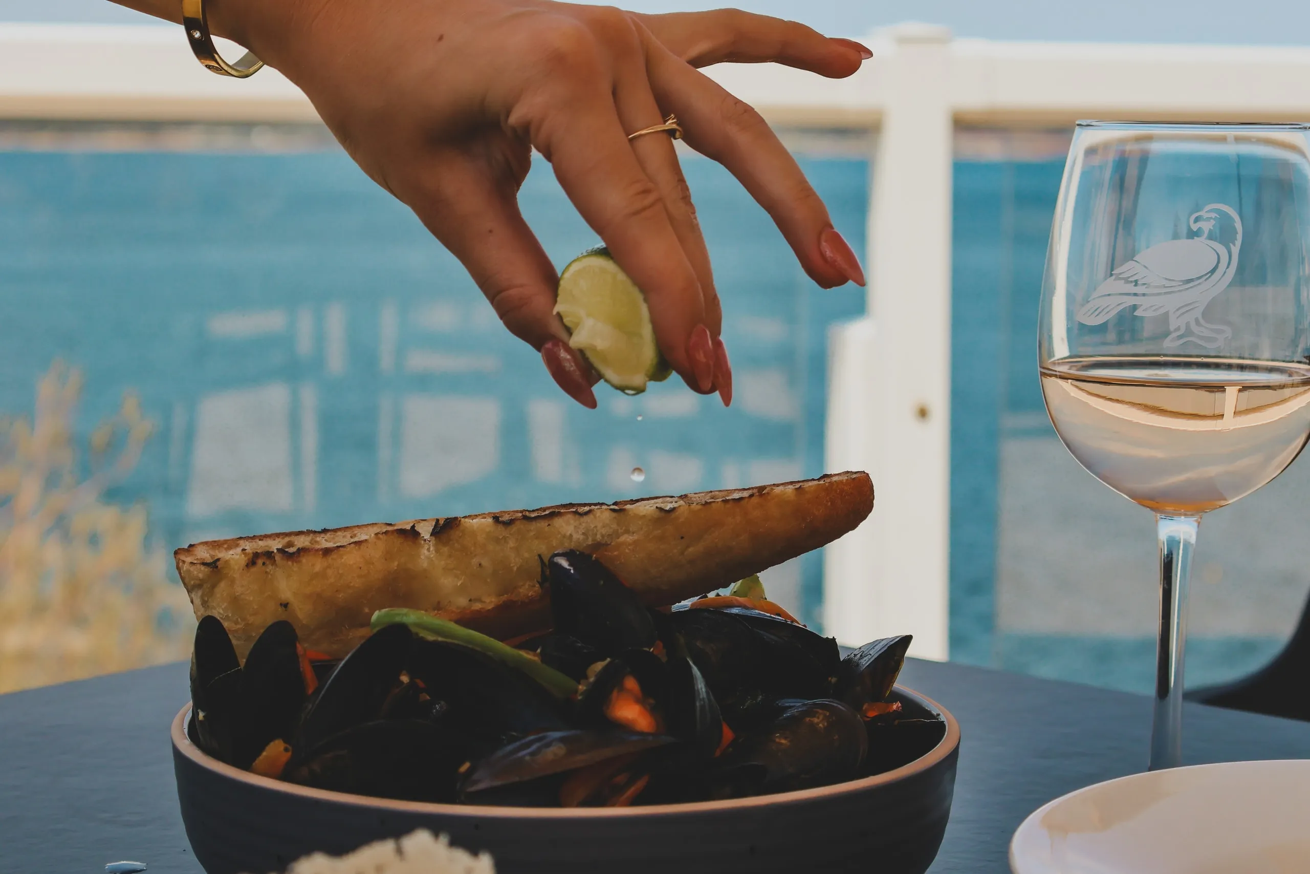 A hand squeezing lime over mussels beside a glass of wine.