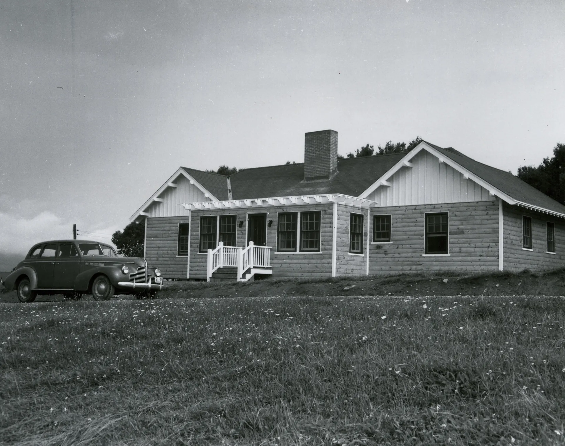 A historical photograph of a white wooden residence with a vintage automobile parked on a daisy-covered lawn.