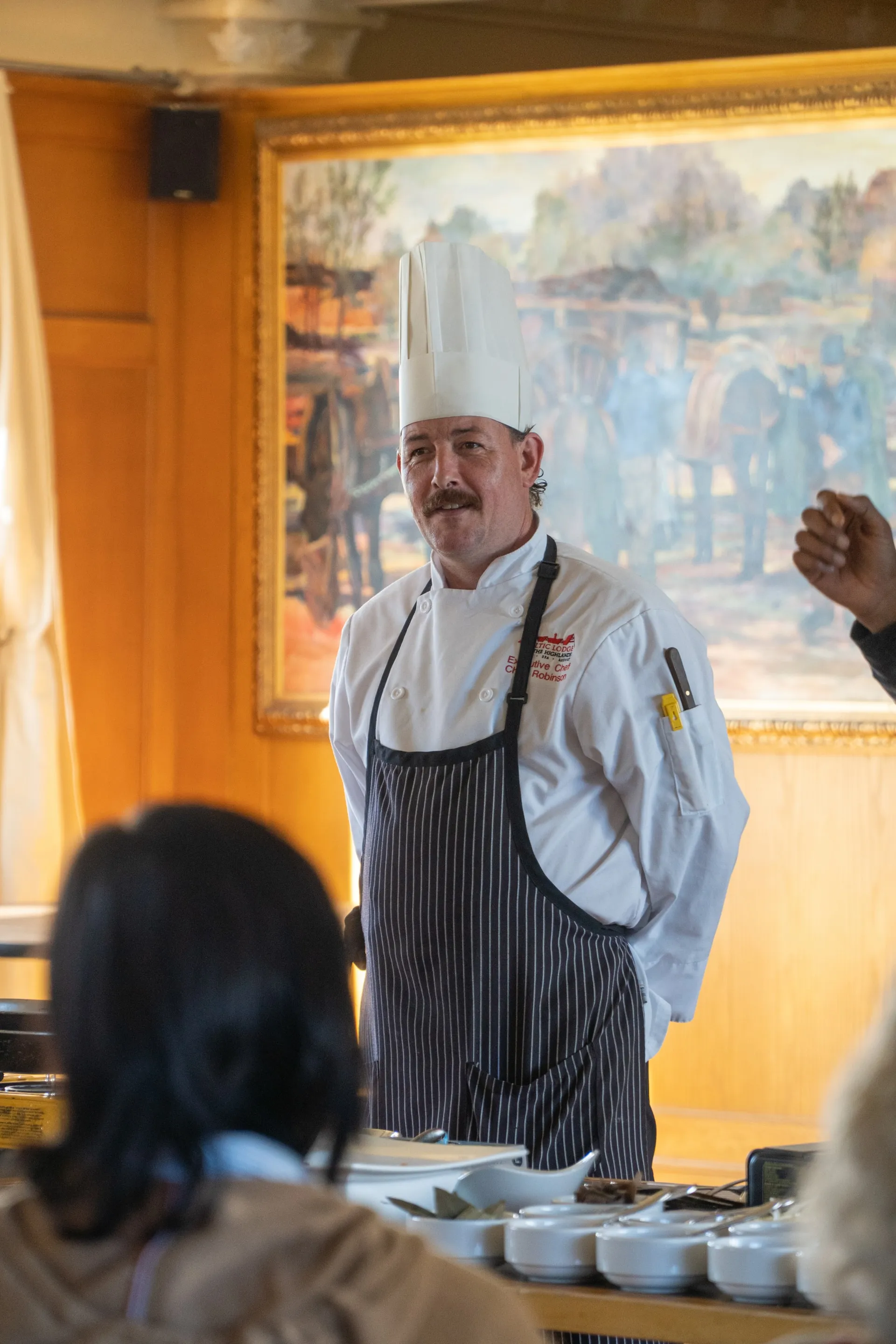 A chef in a white toque and striped apron speaking with the dining team in front of a landscape painting.