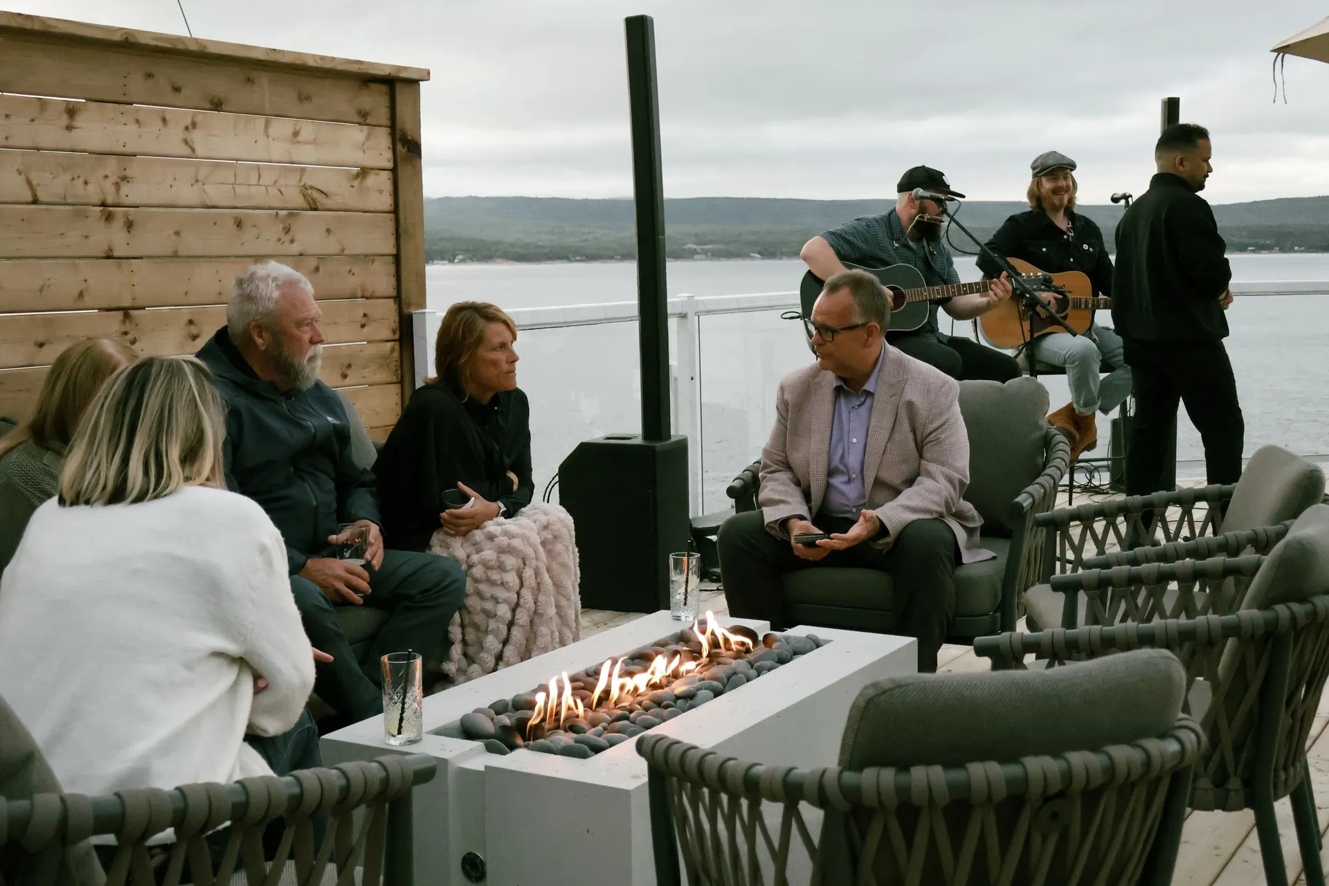 Guests gathered around a fire pit on the terrace while musicians play, with the ocean and hills behind them.
