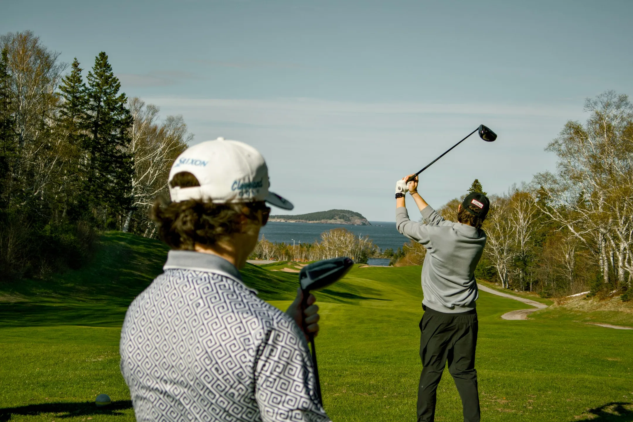 Two golfers on a Highlands Links fairway above the Atlantic, one mid-swing, with an island on the horizon.