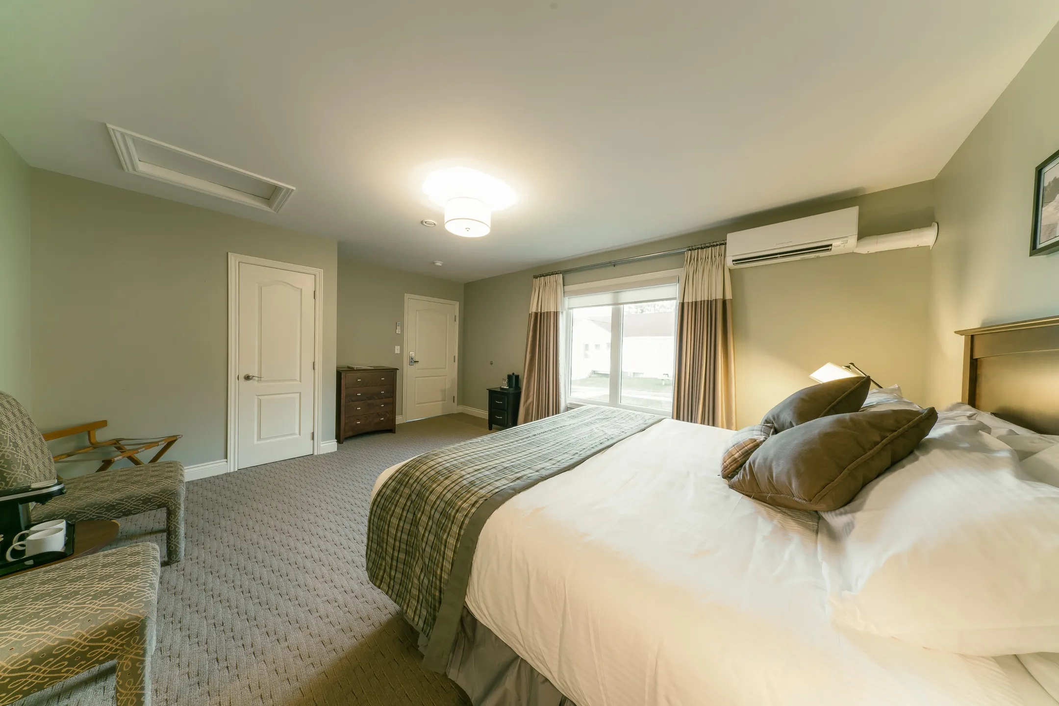 A bedroom with white linens, a patterned throw, and a window dressed with neutral curtains.