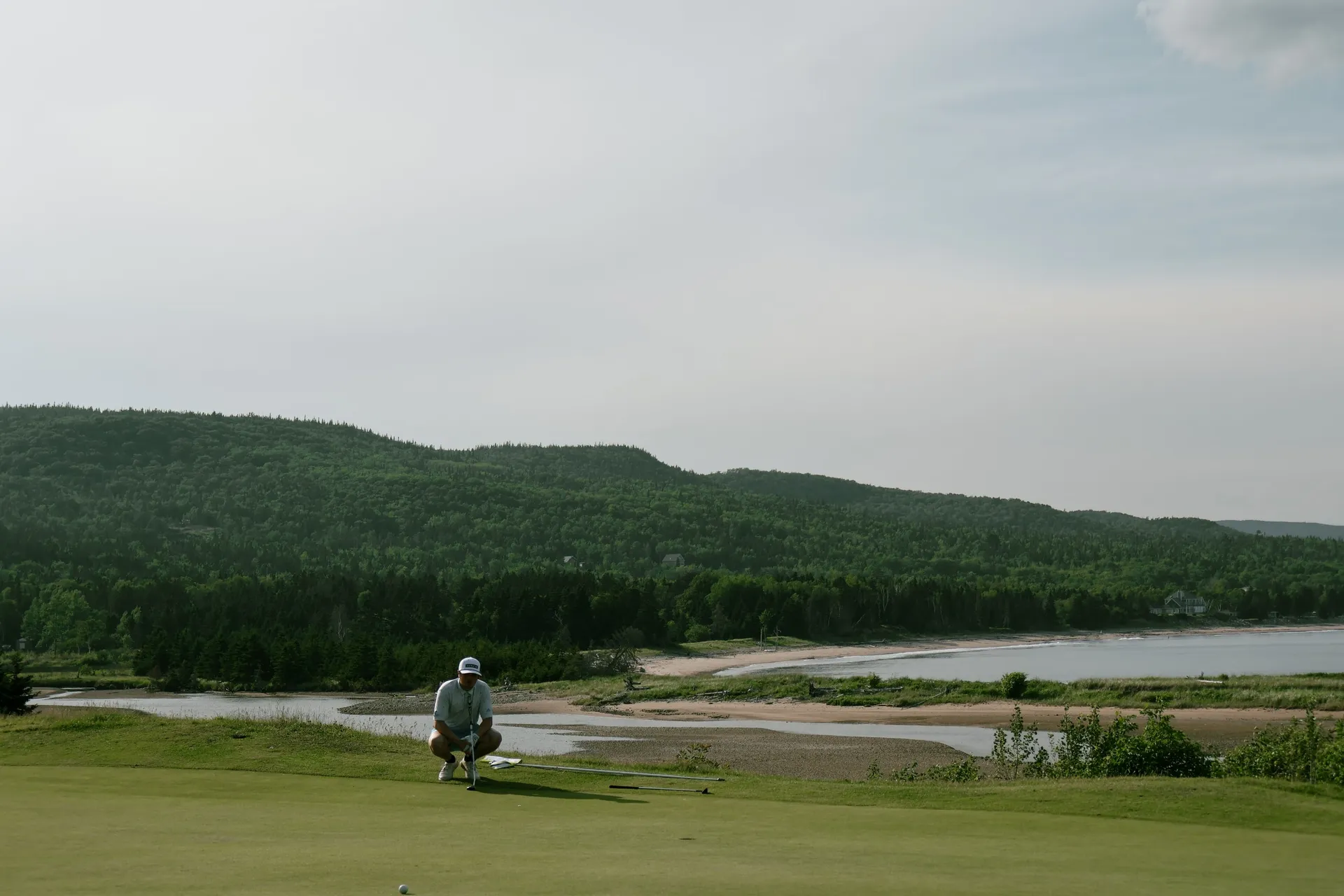 A golfer lining up a putt on a green above a river and forested hills.