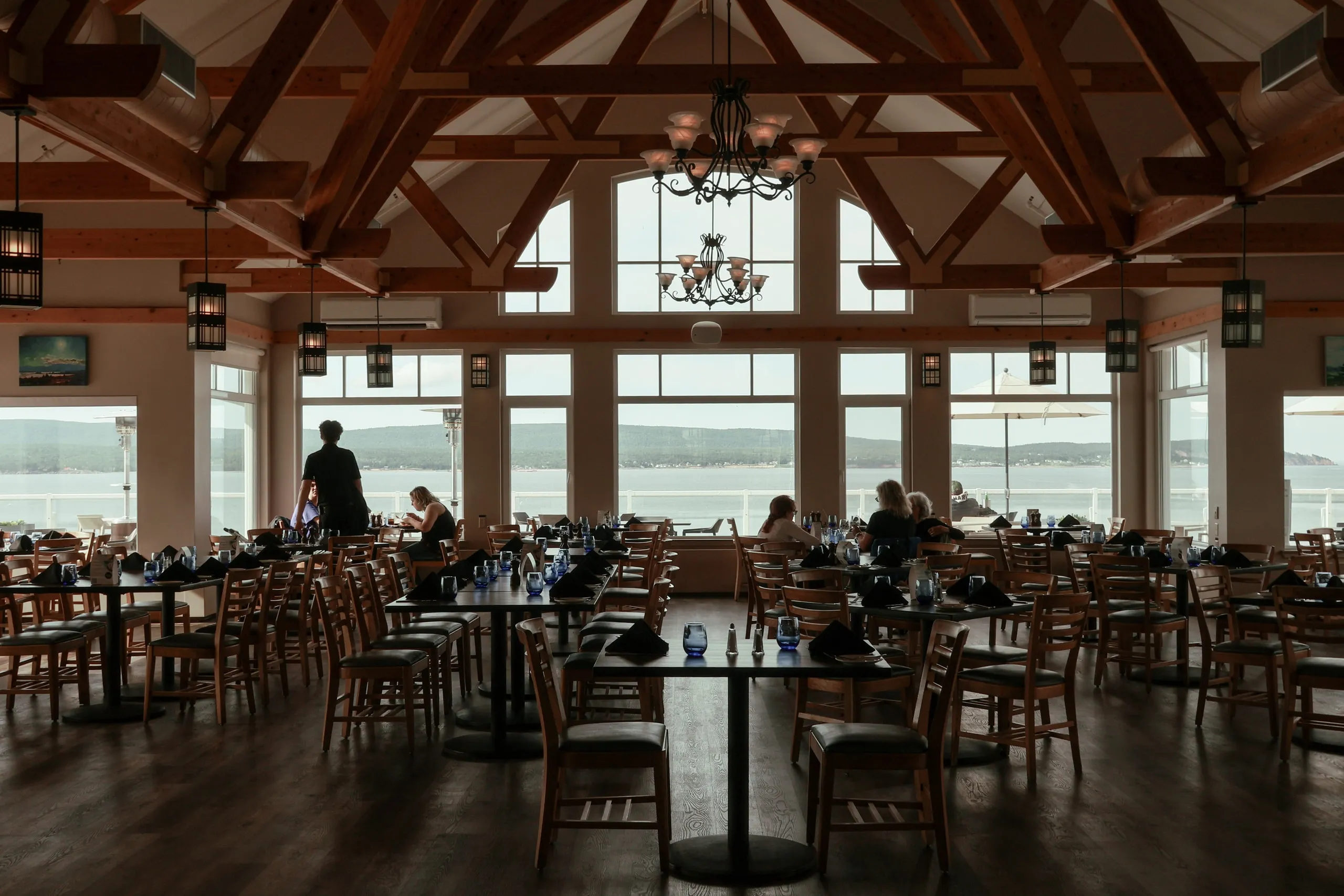 The dining room with wooden beams, set tables, and windows onto the water.