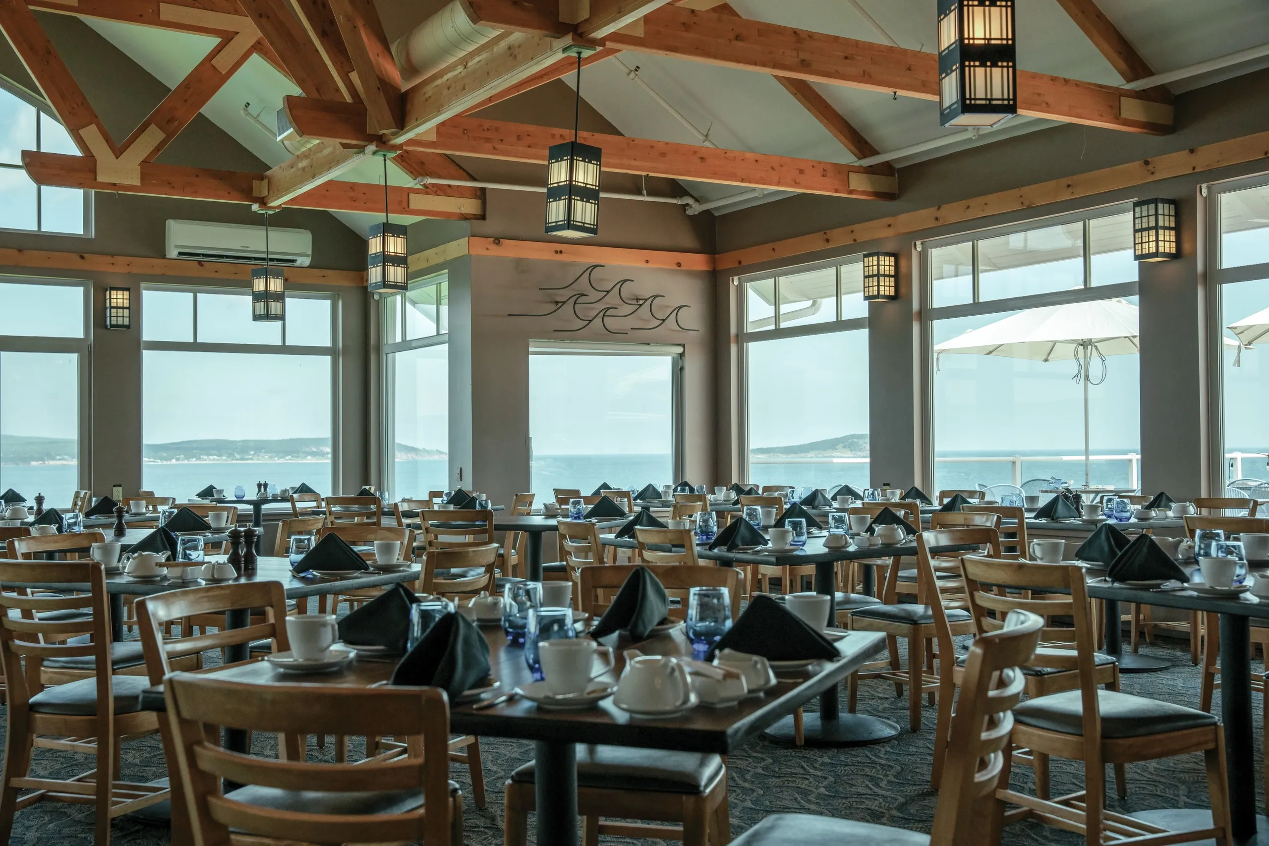 Dining tables set with glassware beneath wooden beams and ocean windows.