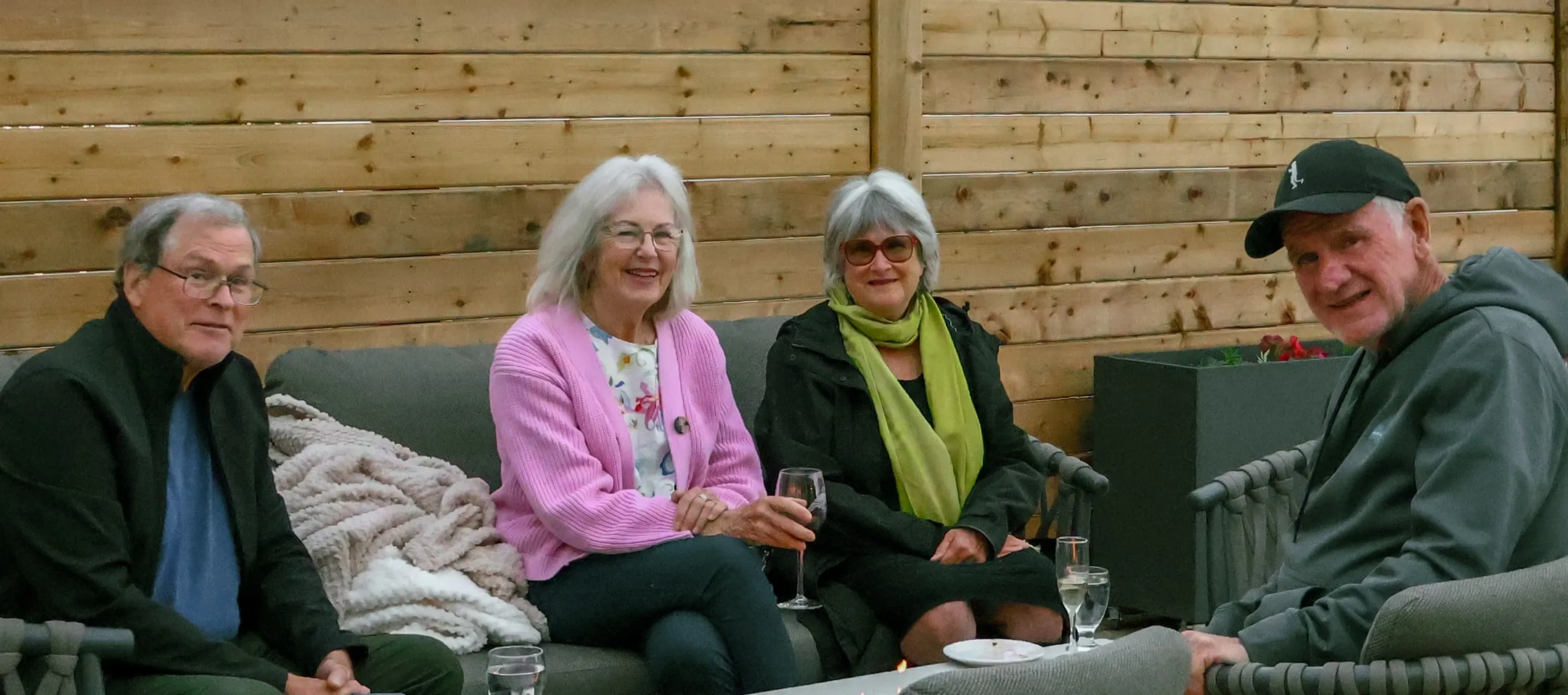 Four guests seated together on an outdoor sofa against a wood wall, with drinks on the table in front of them.