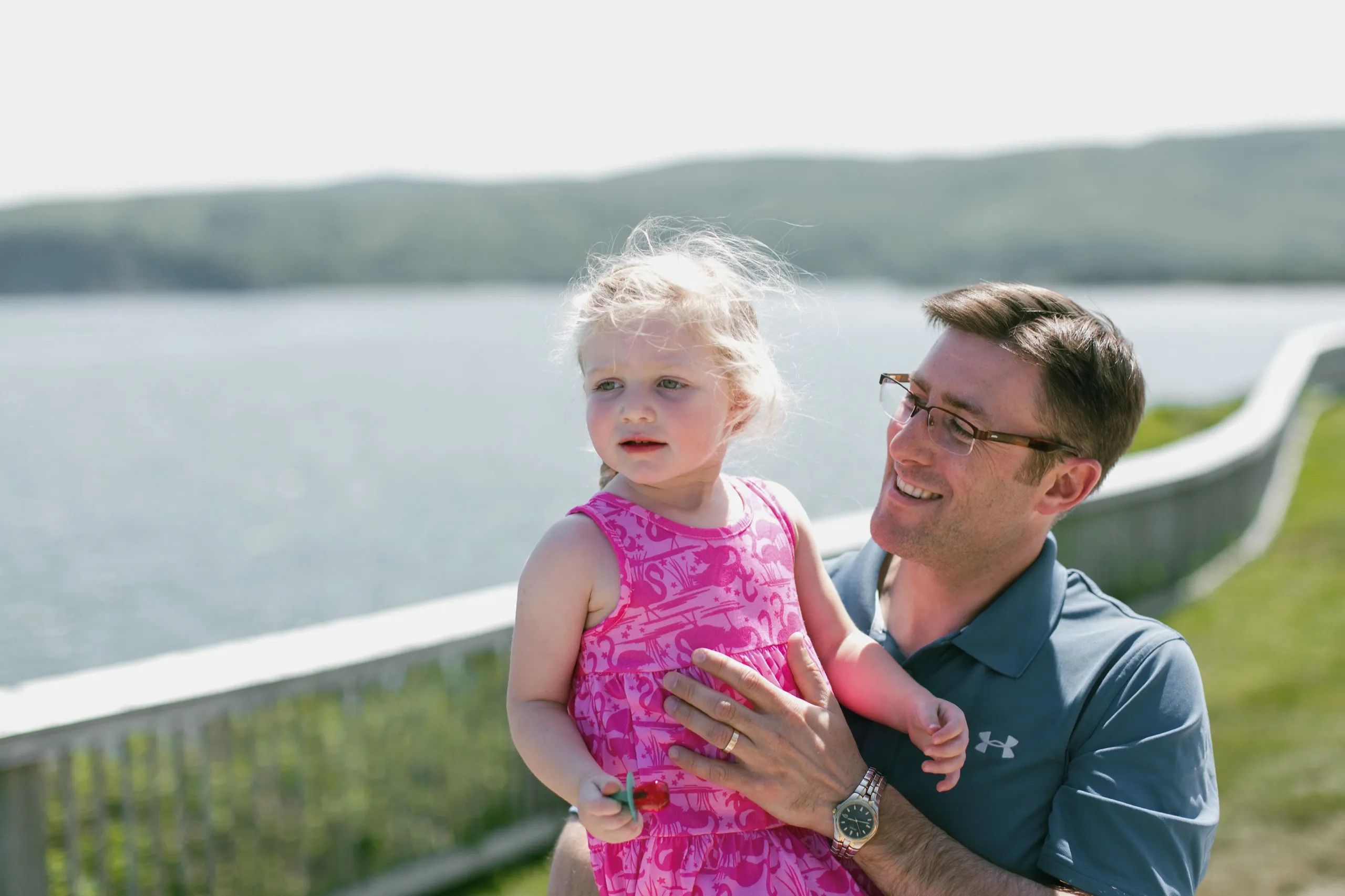 A father holding his young daughter outdoors with water and green hills in the background.