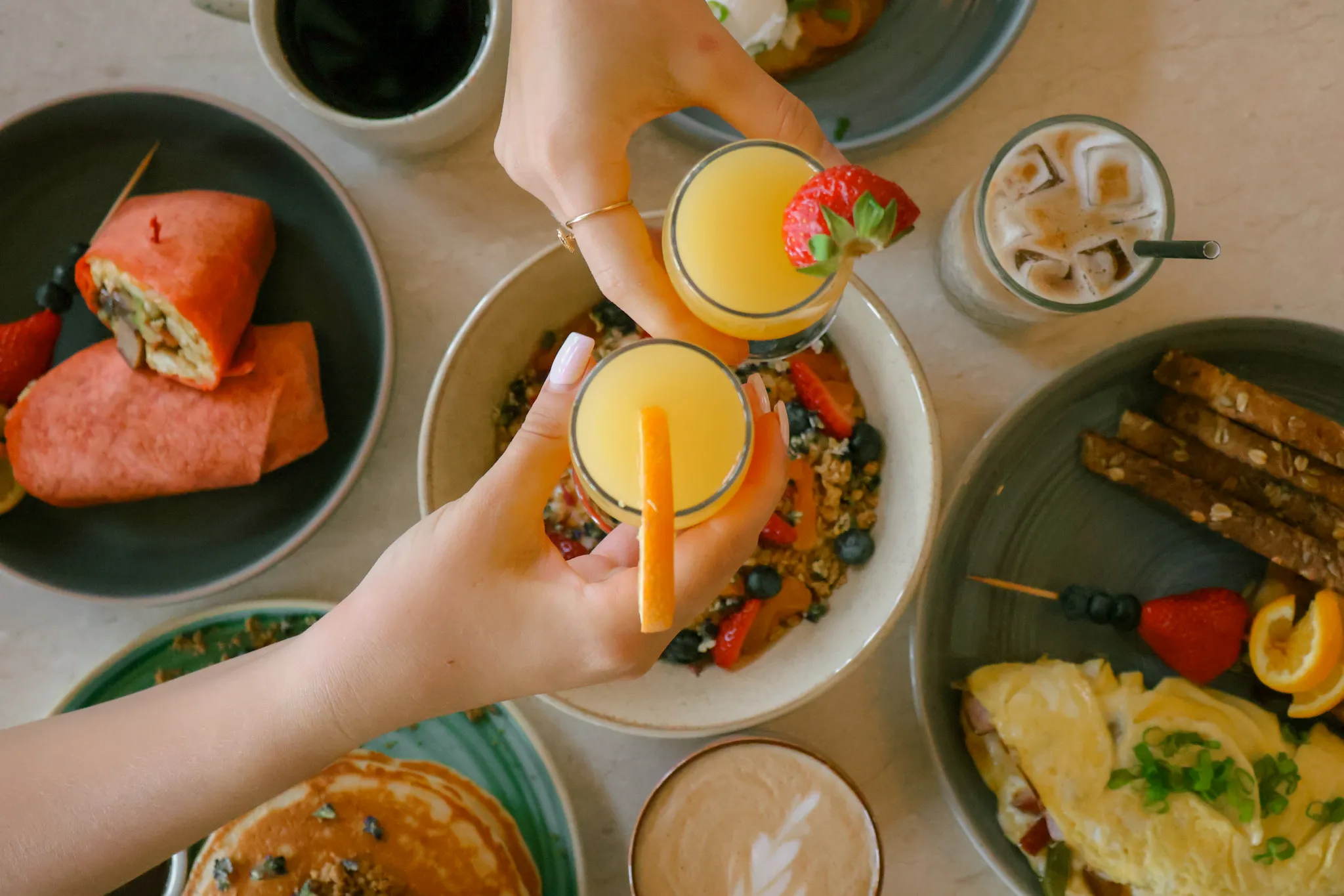 Overhead view of a breakfast table, with two hands toasting glasses of orange juice over granola, pancakes, and an omelette.