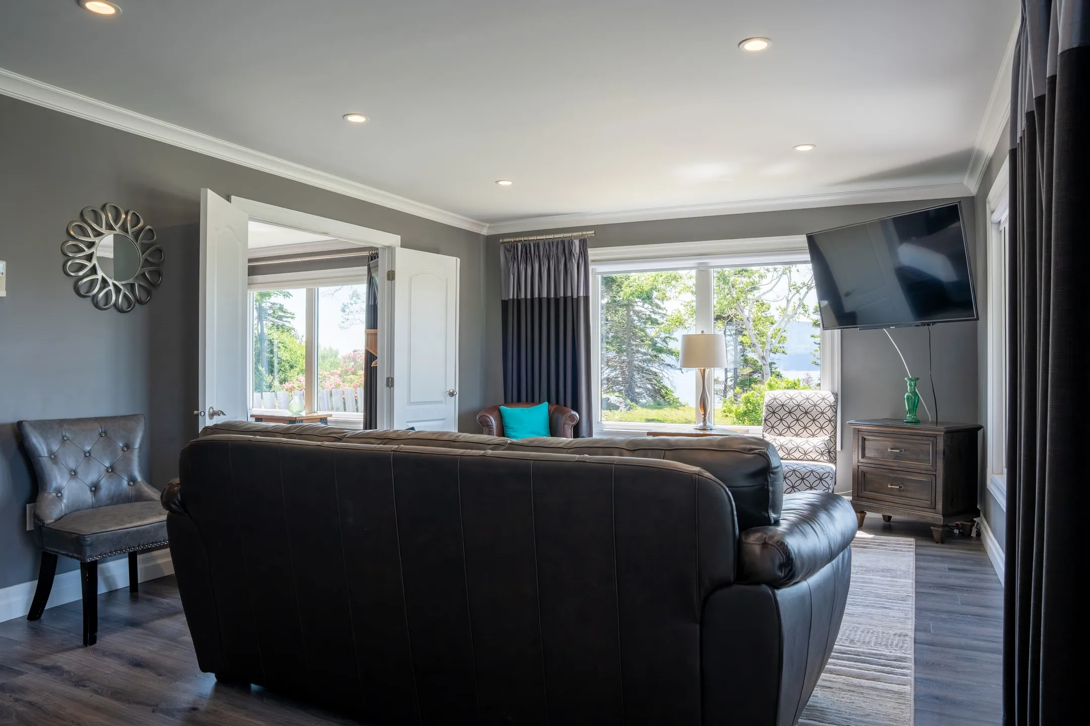 A living room with a dark leather sofa, armchairs, and windows opening onto trees and the grounds.