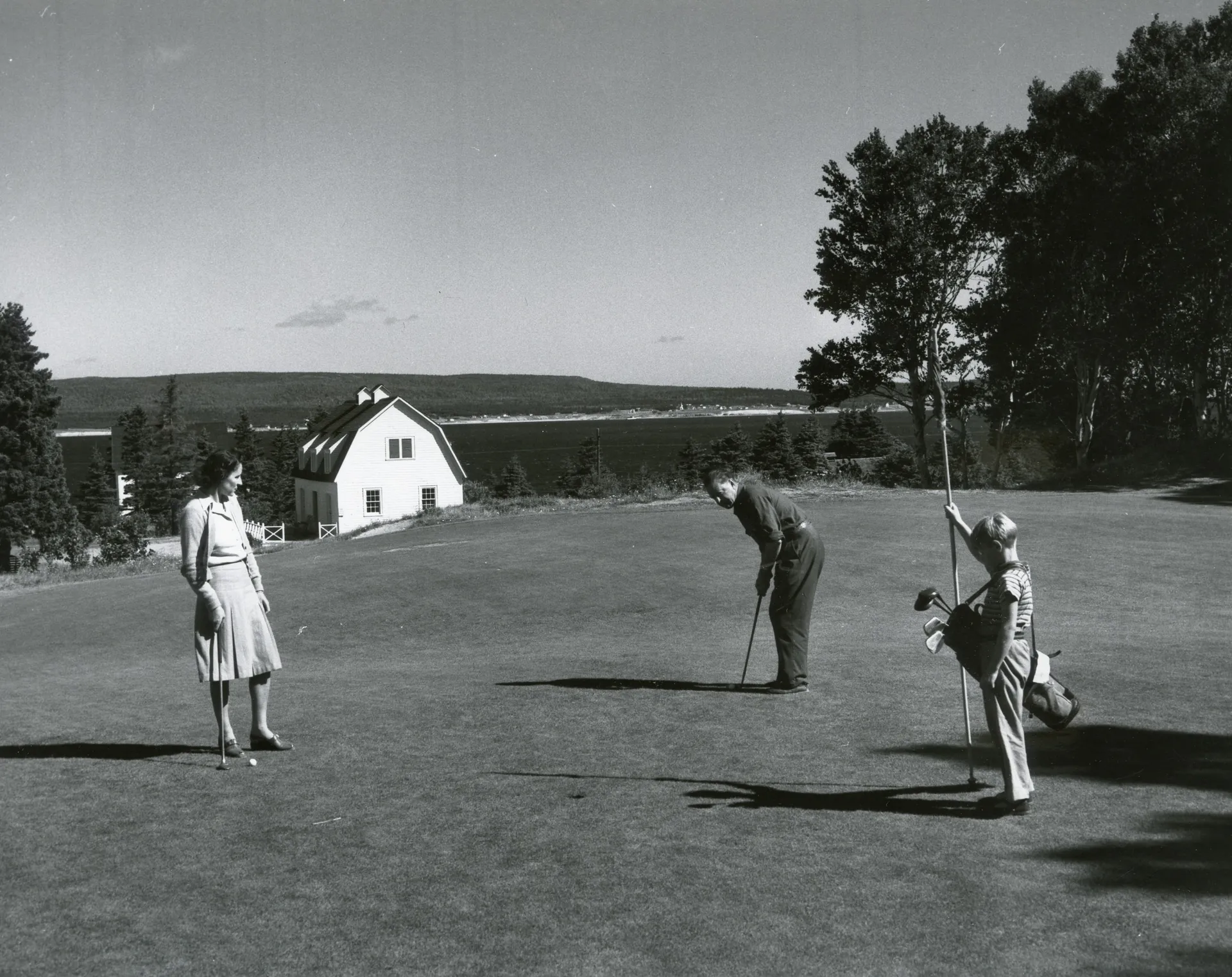 Mid-century golfers on the fairway at Cape Breton Highlands Links, with a white cottage and the coast behind them.