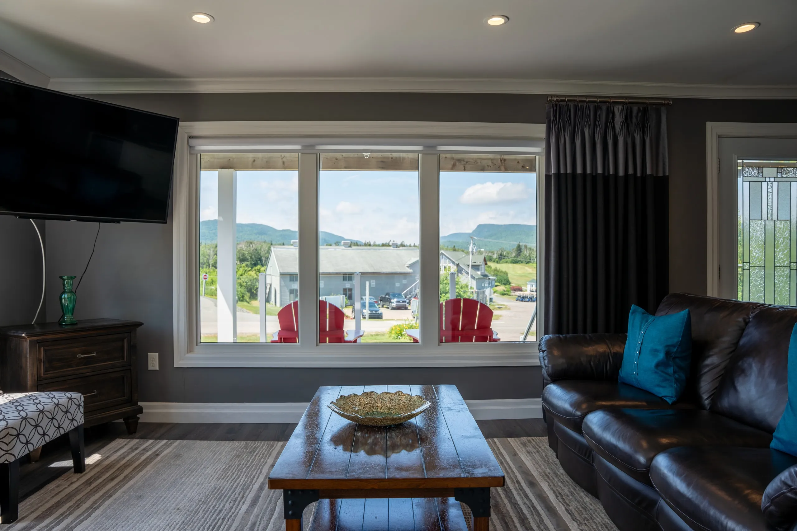 A living room looking out through a large window to red Adirondack chairs on the deck and green hills beyond.